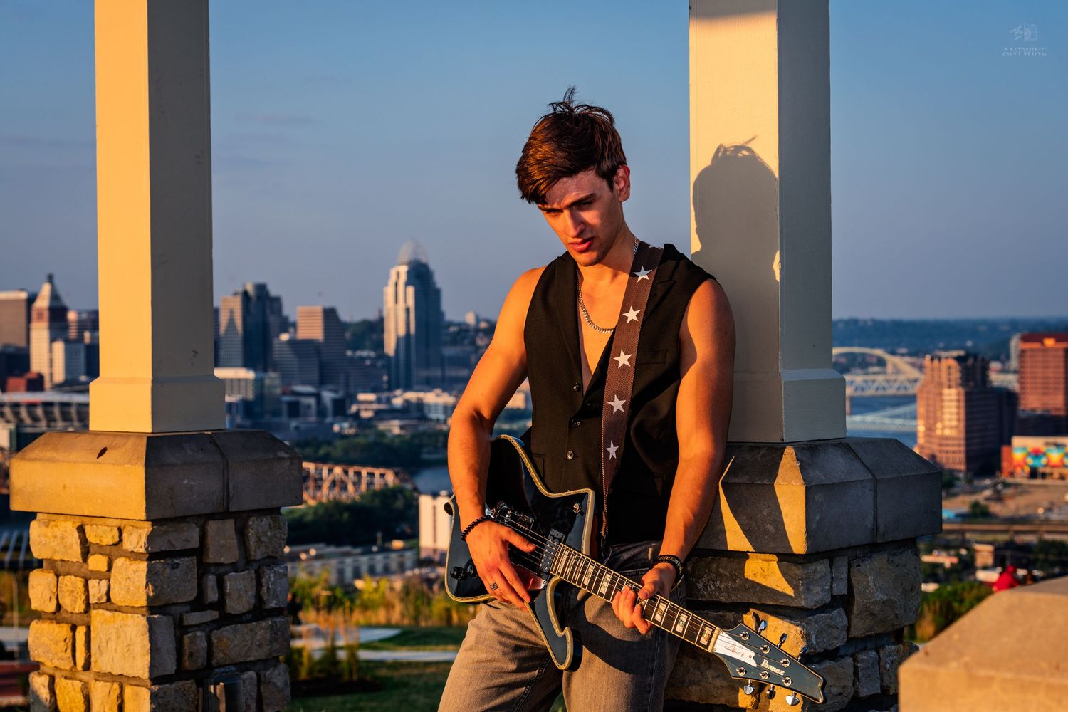 Young male guitarist playing electric guitar on rooftop at sunset with city skyline in background.