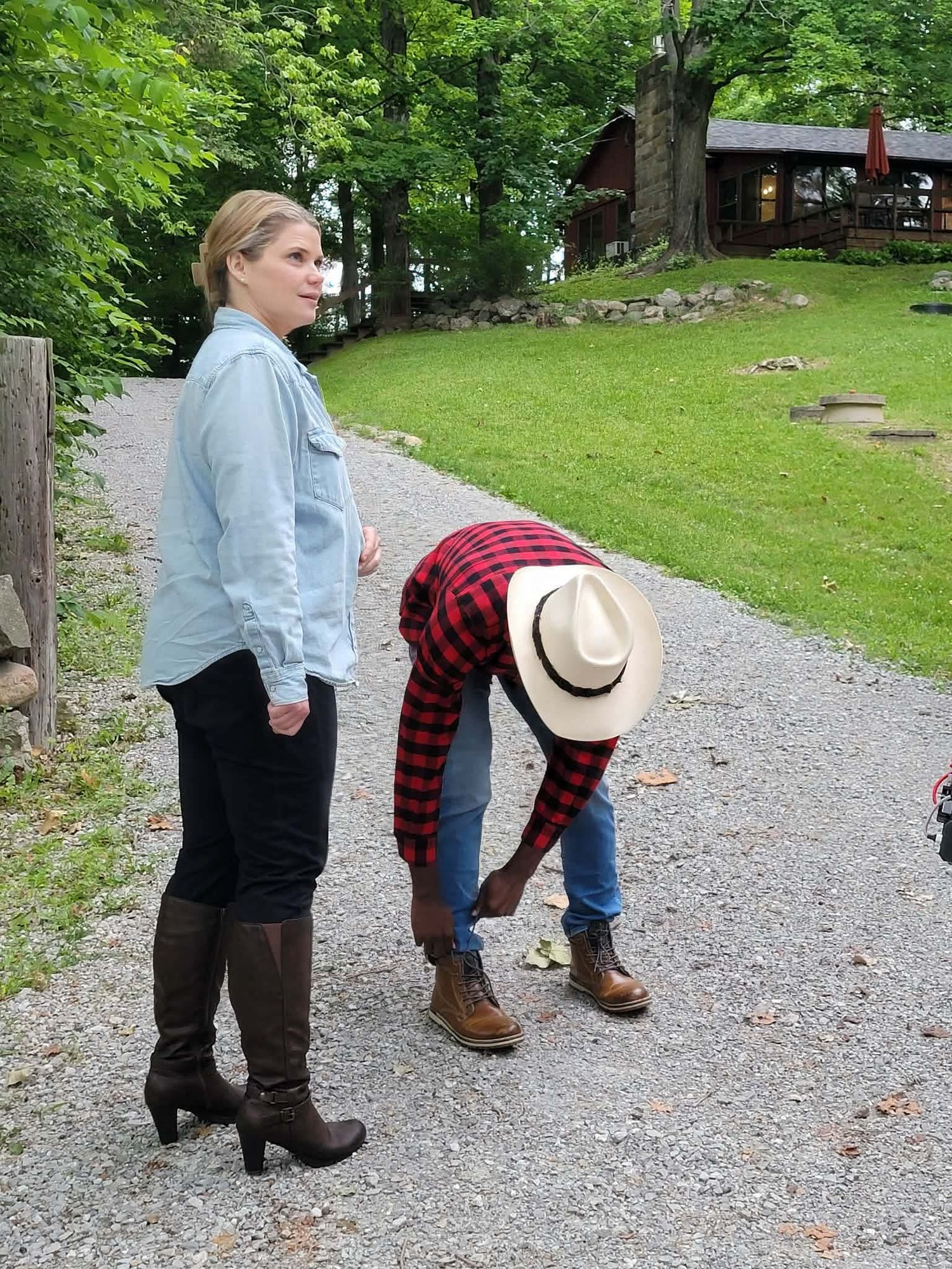 Woman in blue shirt and boots stands on gravel path while man in plaid bends down near rural cabin.