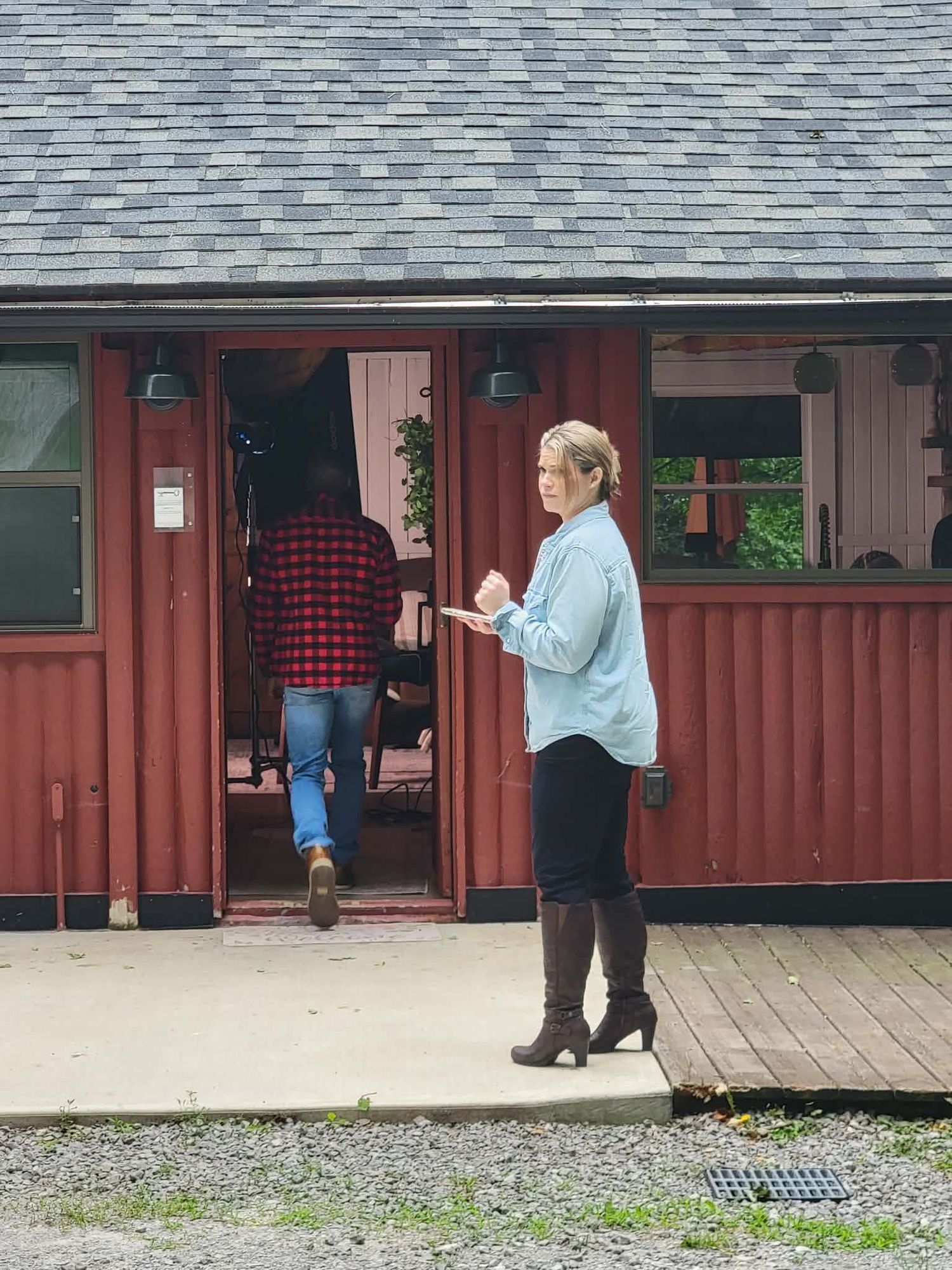 Woman in blue shirt and boots stands outside a rustic red barn building while a man in plaid enters the door.