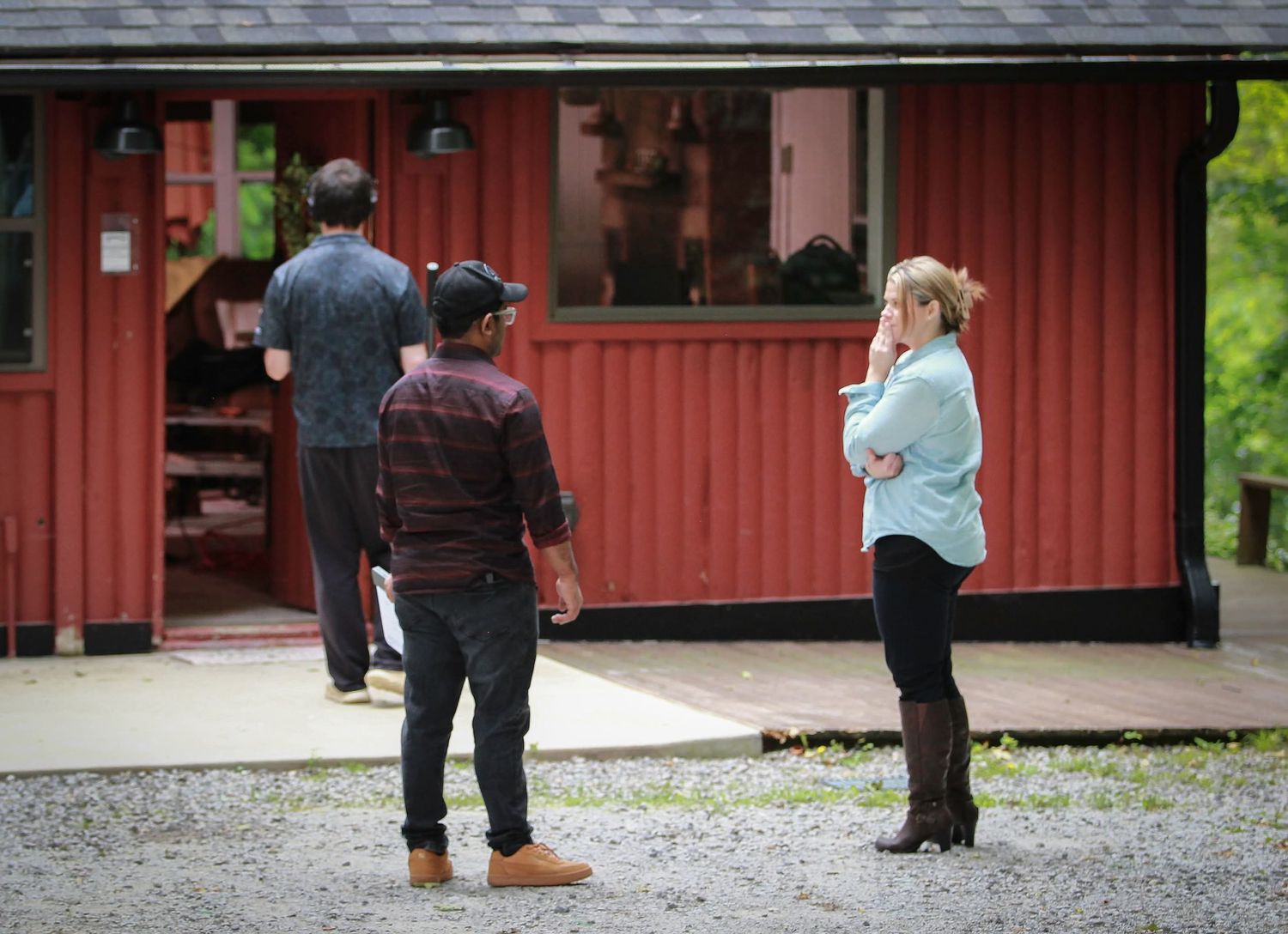 Three people stand outside a rustic red barn building on a gravel lot surrounded by lush green trees.