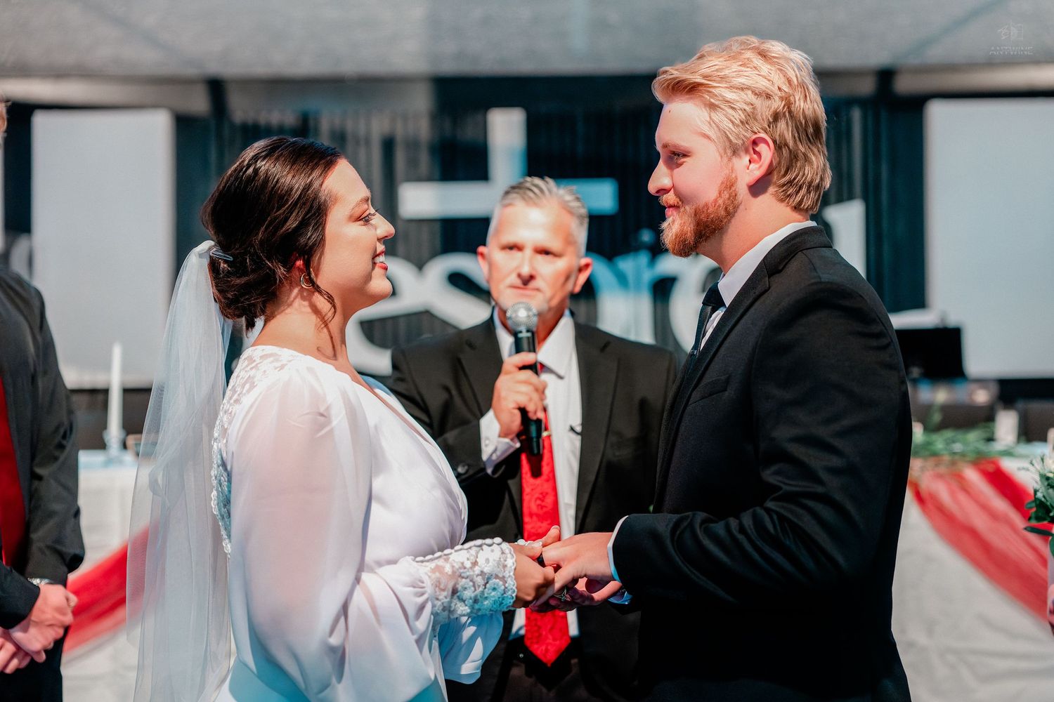 Bride and groom exchange vows at wedding ceremony with officiant holding microphone in background.