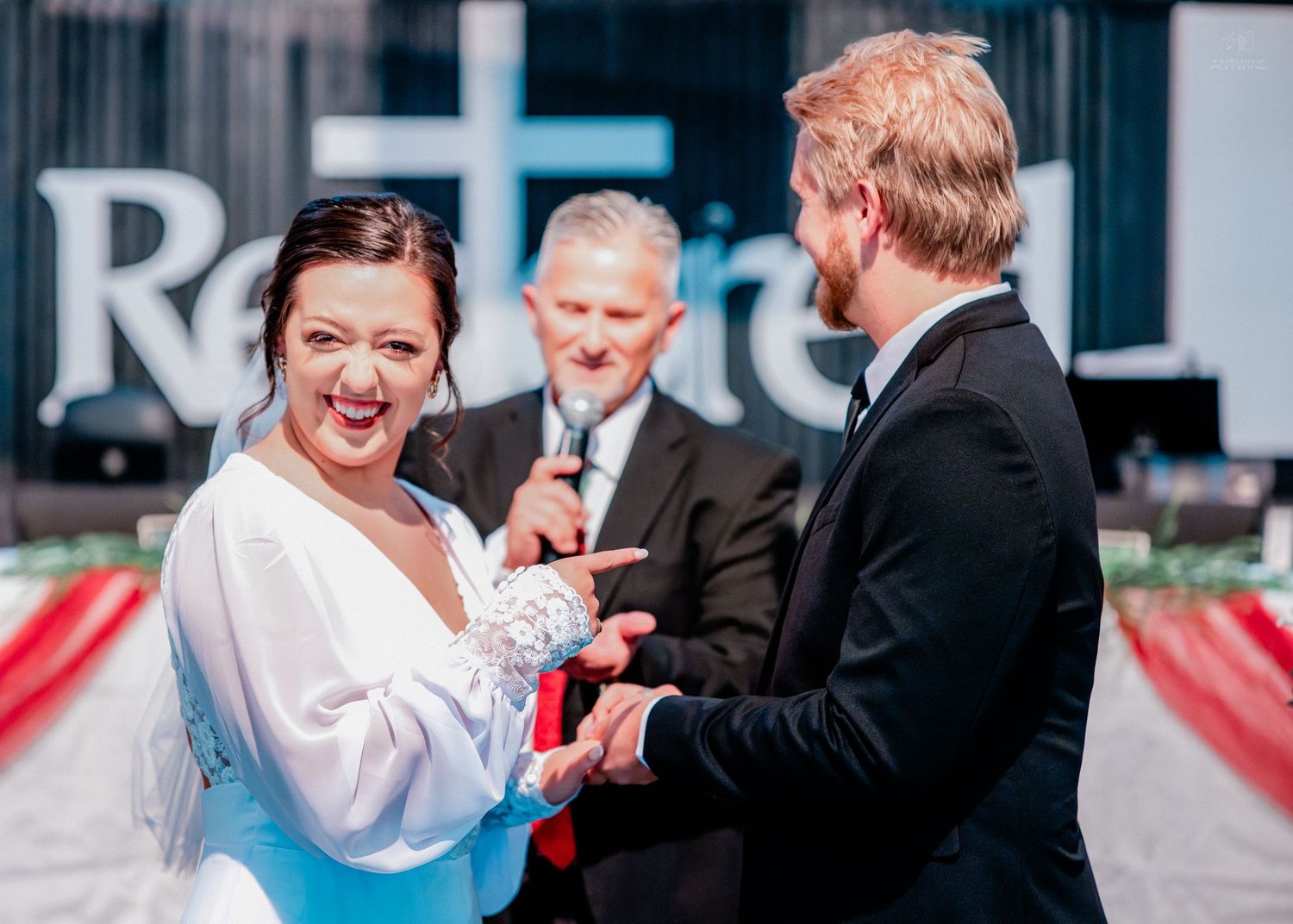Bride and groom exchanging vows at wedding ceremony with officiant in background near red decor.