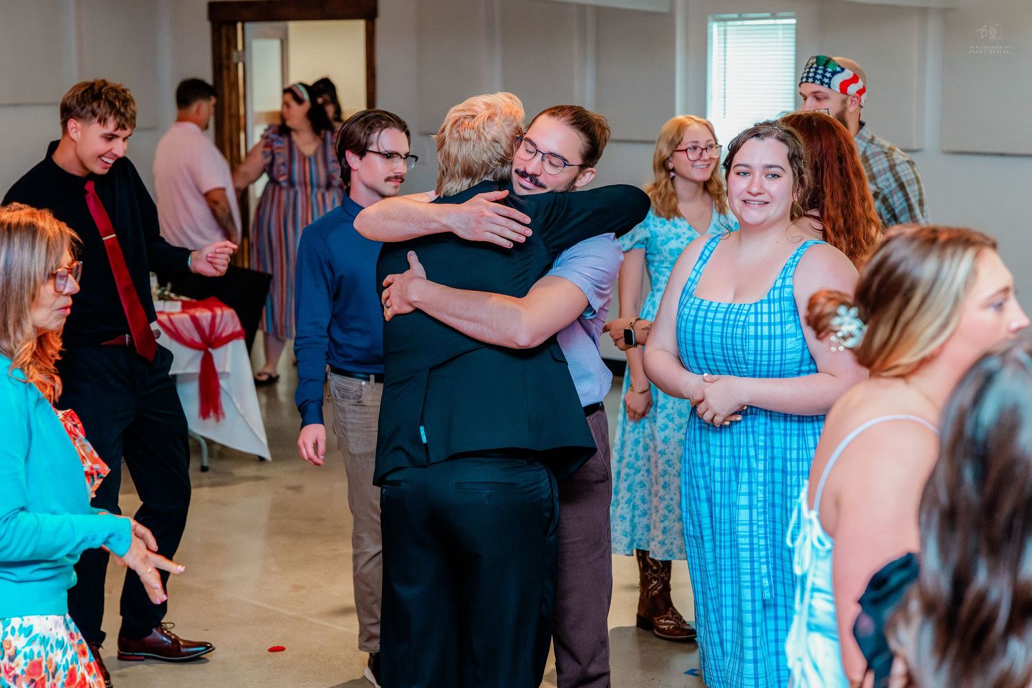 Guests sharing warm hugs and celebrating at a festive indoor event with others mingling in the background.