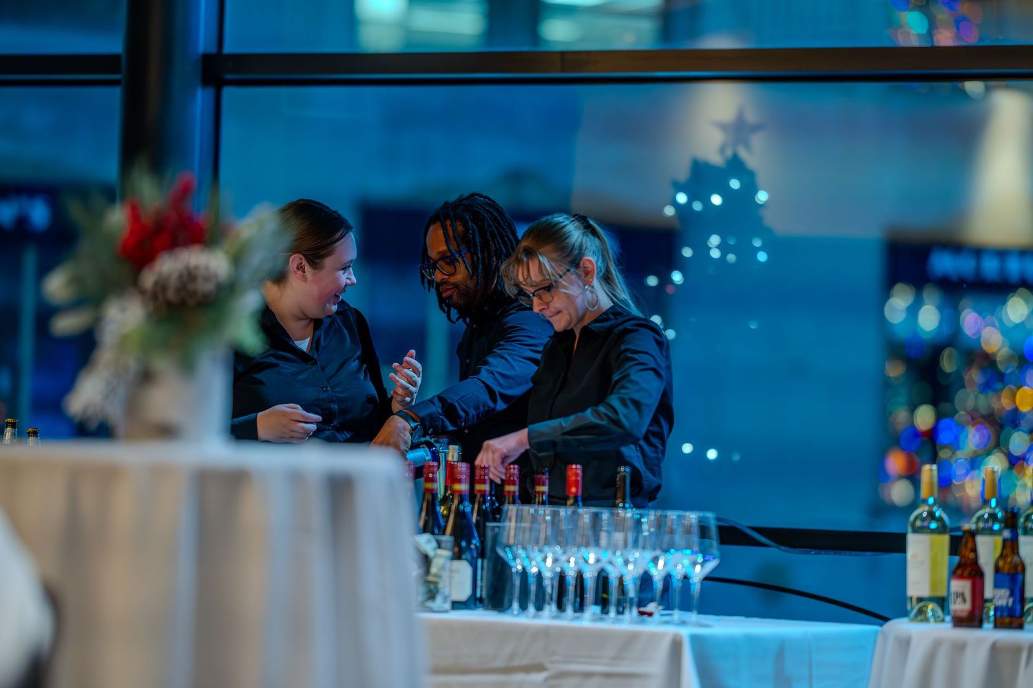 Three bartenders prepare drinks at a festive holiday event with champagne glasses and bottles on a lit table.