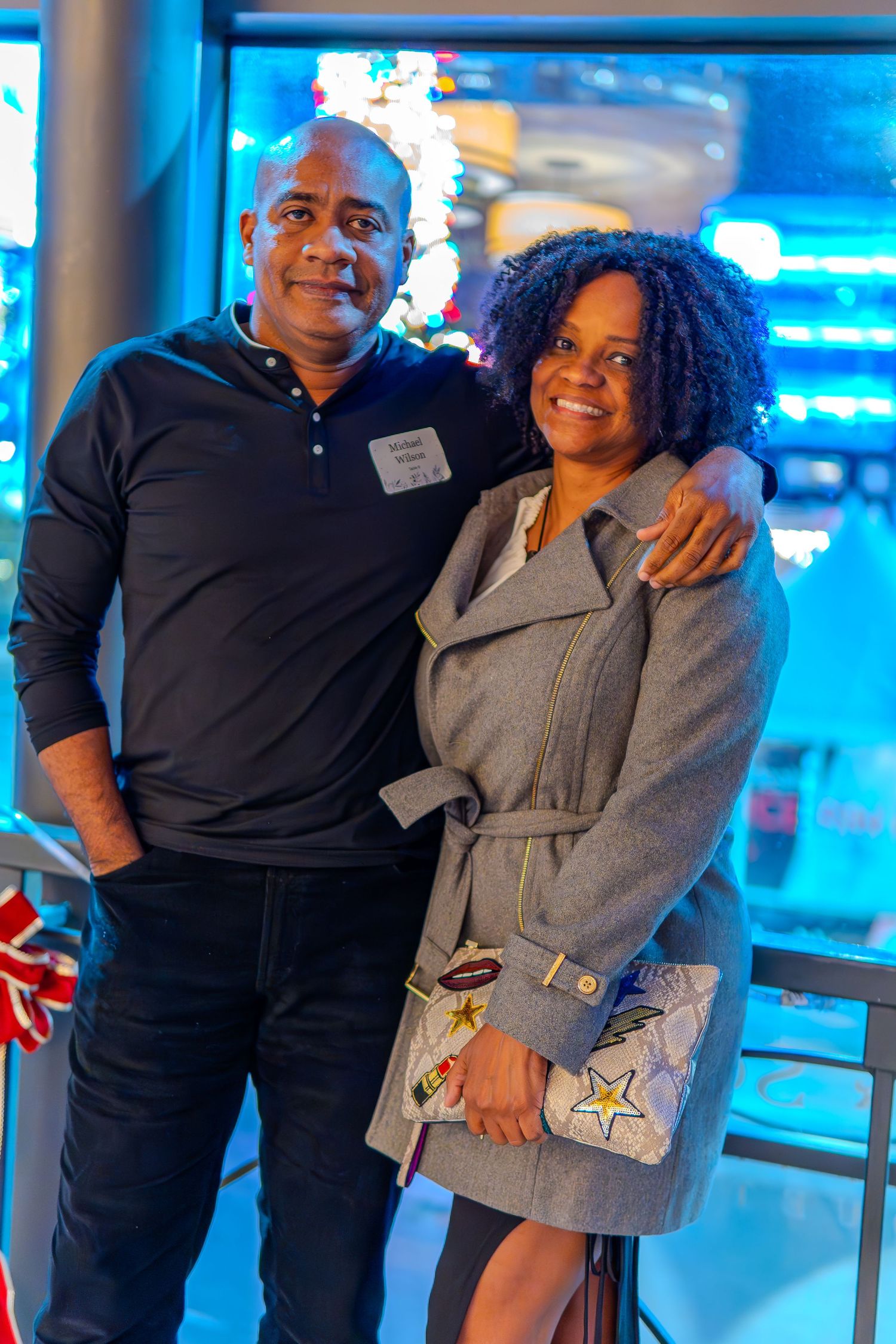 A smiling couple poses together at a festive holiday party with a Christmas tree glowing in the background.