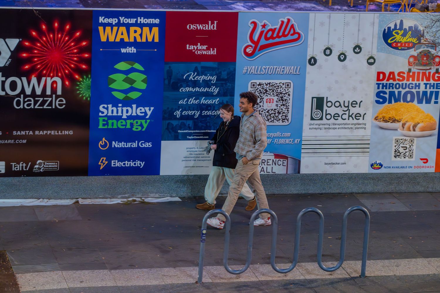 Two people walk past a colorful advertising billboard wall featuring Shipley Energy and local business signs on a city sidewalk.