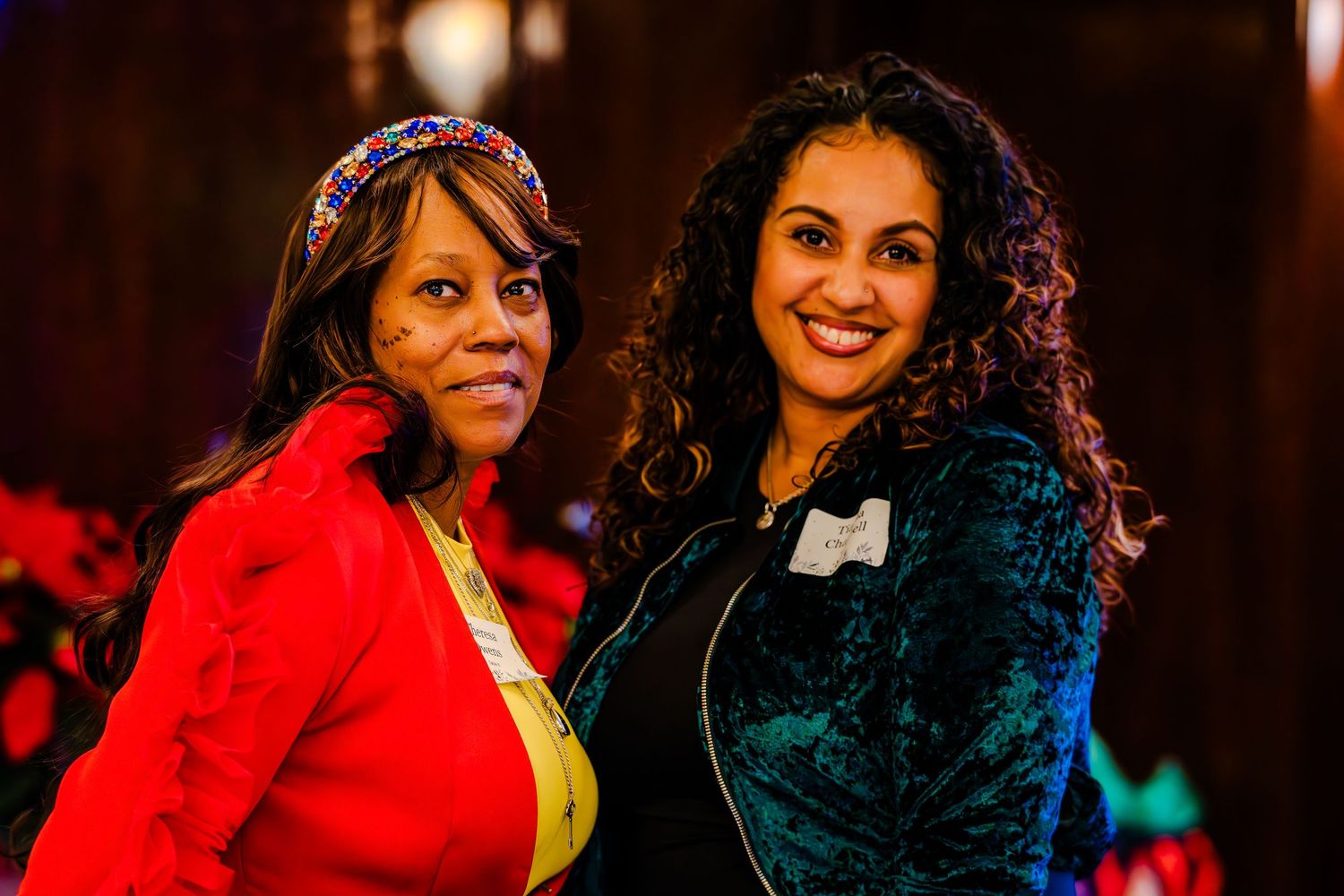 Two smiling women posing together at a festive holiday event, one in red and one in teal jacket.