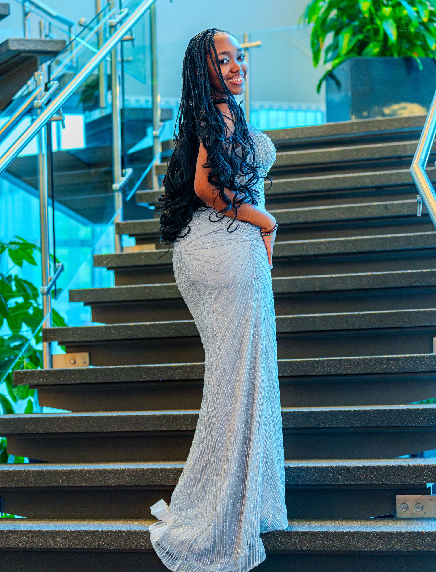 Woman in fitted silver sequin gown with long braids posing on stairs.