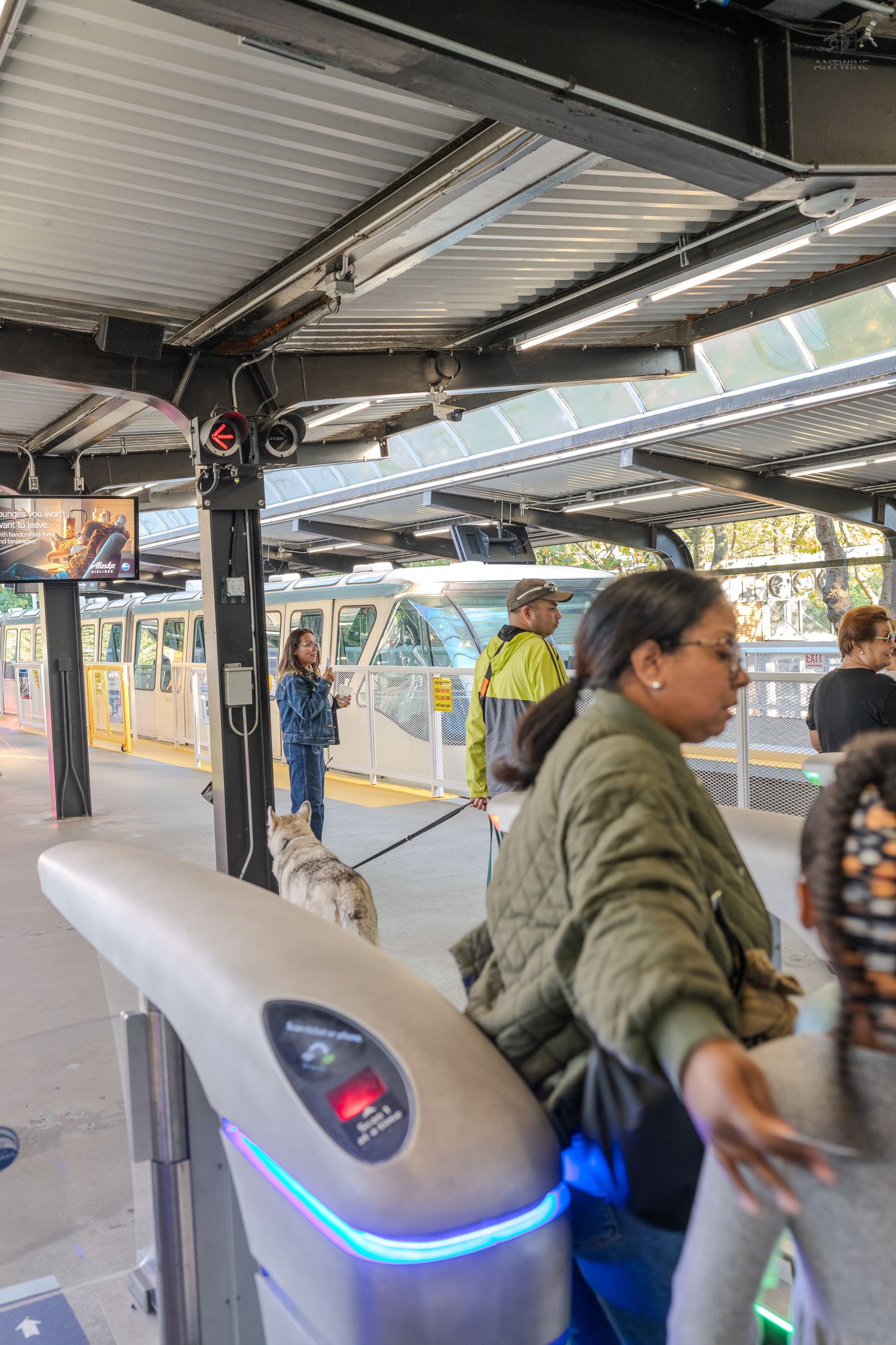 Woman taps fare gate at a Seattle monorail station as passengers and a service dog wait nearby.