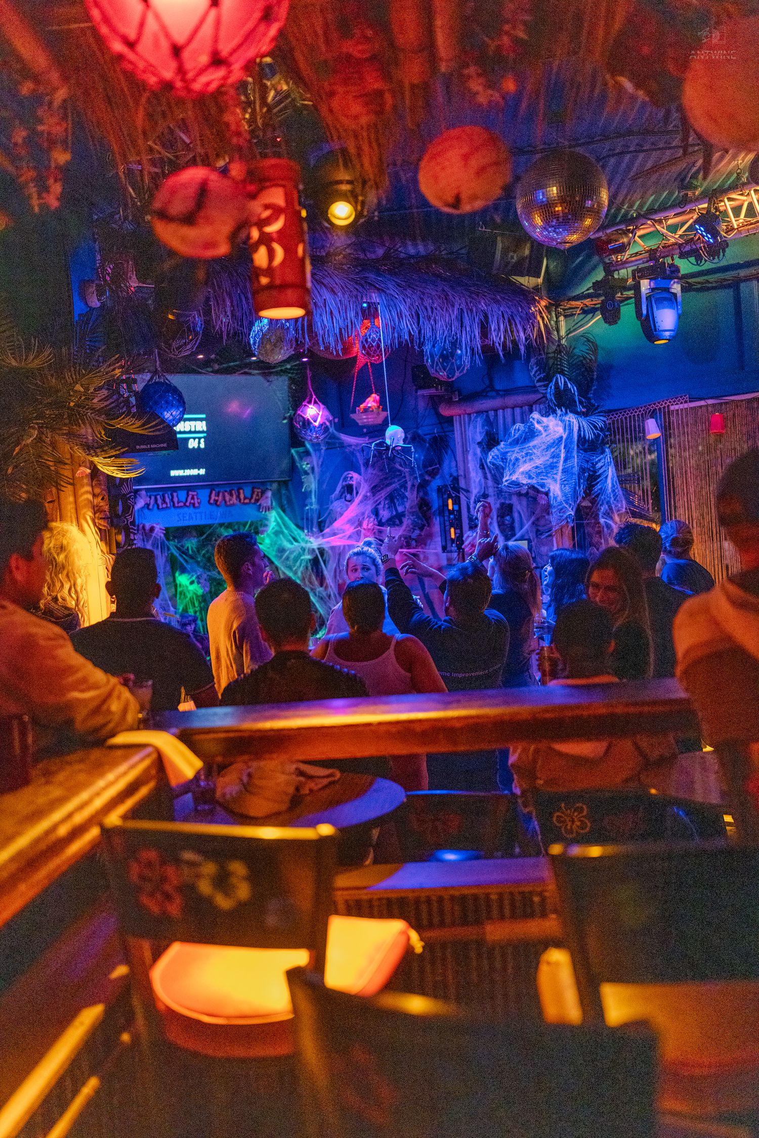 Colorful tiki bar interior with neon lights, tropical decor, and Halloween decorations as guests socialize.