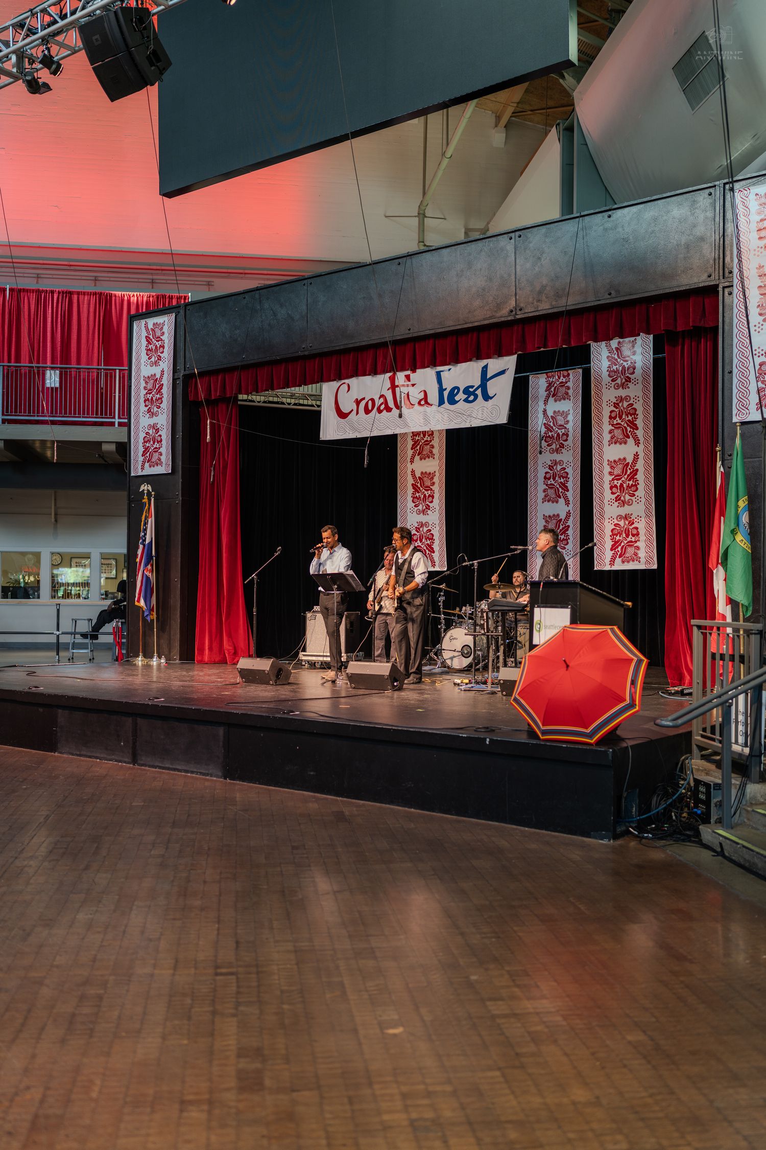 Live band performs on stage at CroatiaFest with red curtains, decorative banners, and a red umbrella in foreground.