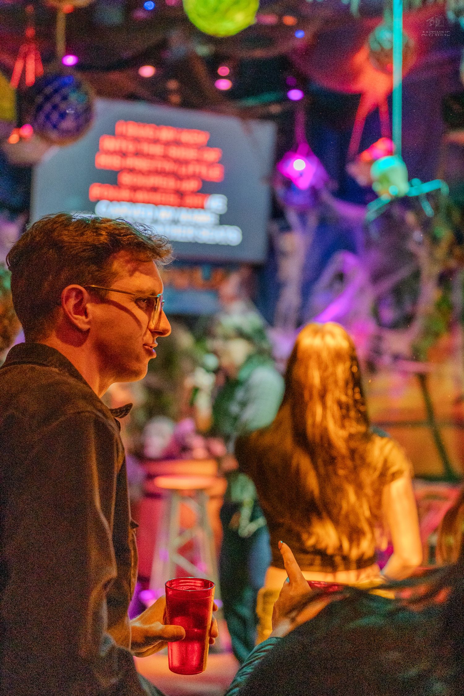 Man holding red cup watching karaoke performance at colorfully decorated Halloween themed bar.