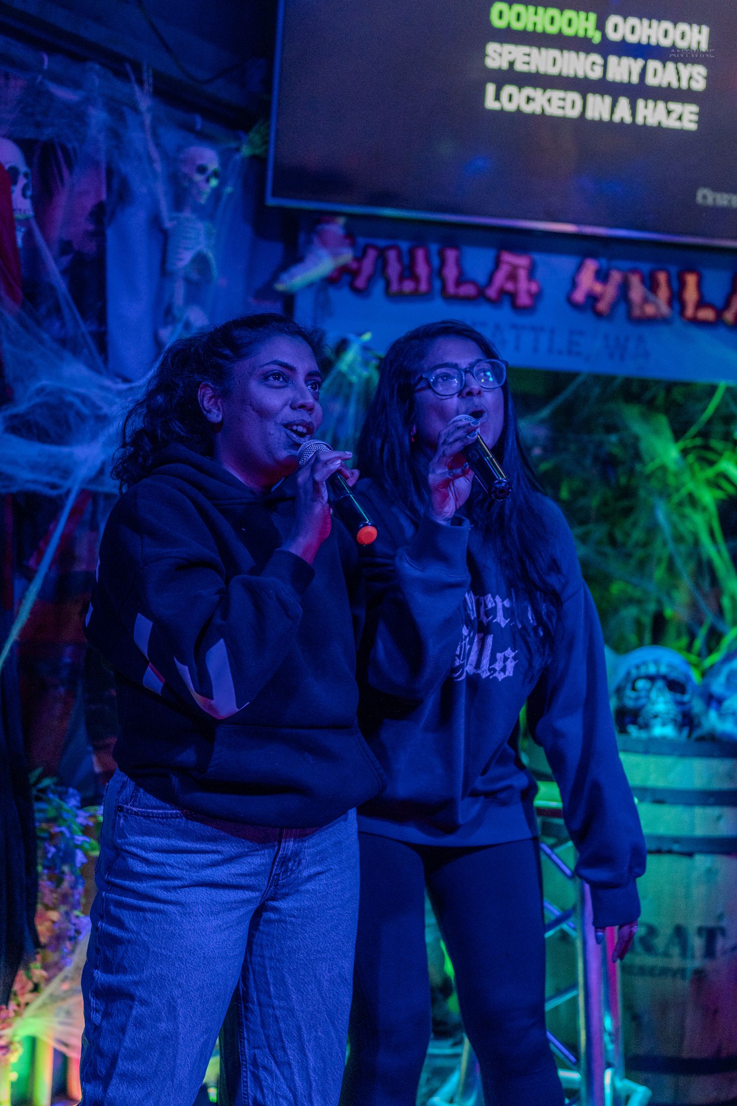 Two women singing karaoke duet on Halloween-decorated stage with microphones at Hula Hula bar.