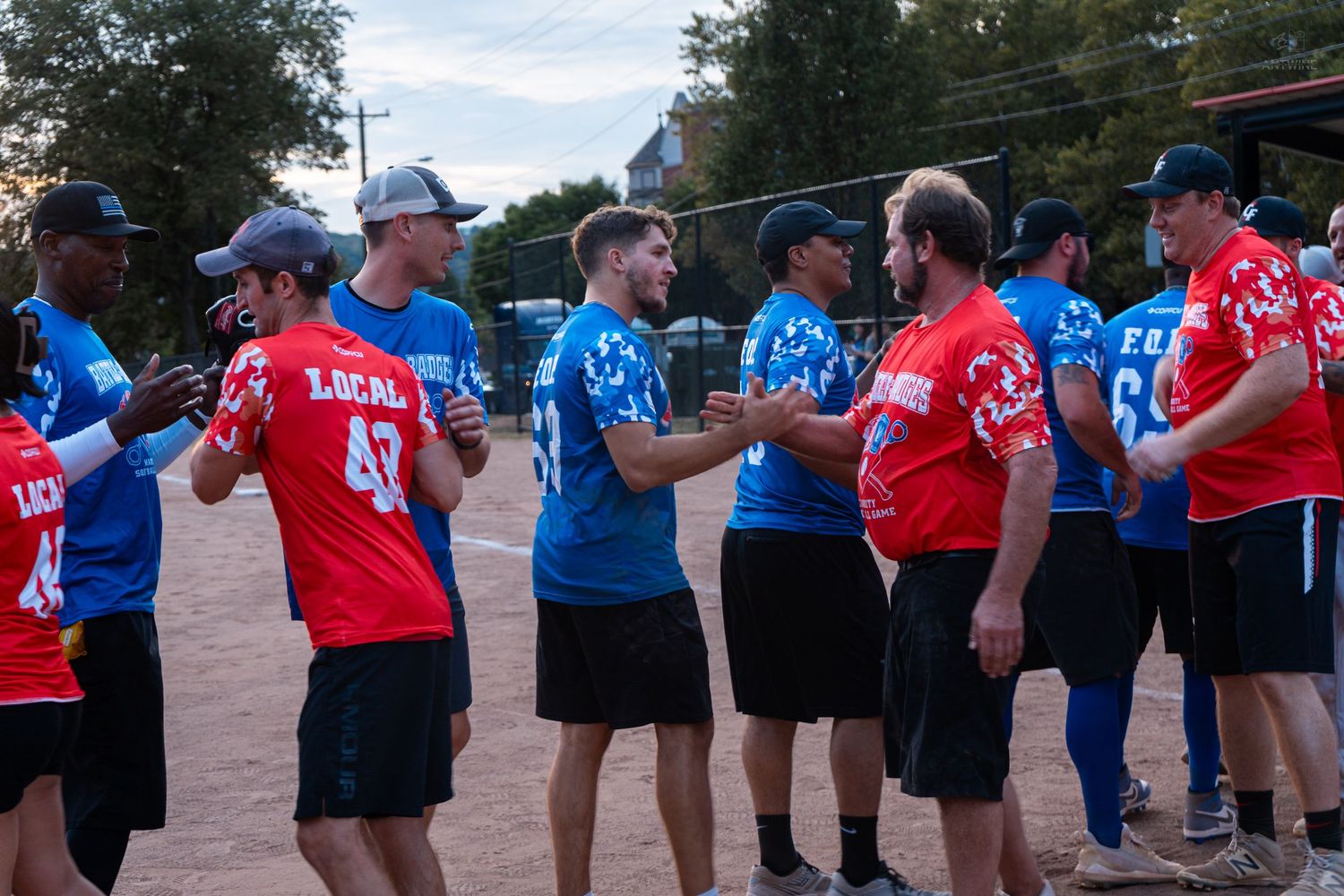 Two softball teams in red and blue jerseys shake hands on a dirt field after a game.