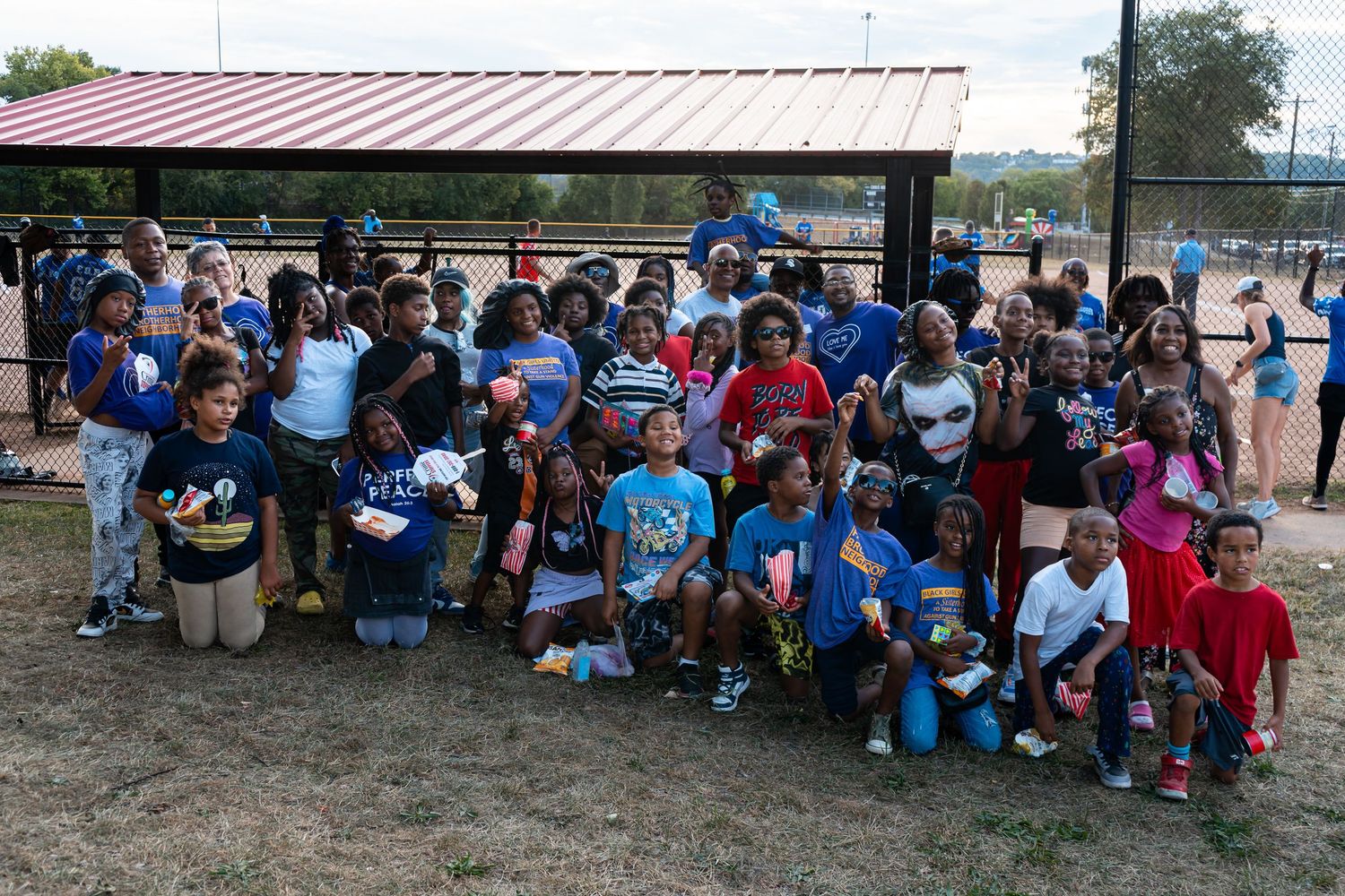 Large group of children and adults pose together smiling at an outdoor community baseball field event.