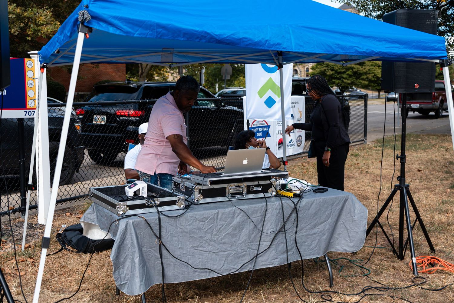 Outdoor DJ setup under blue canopy tent with turntables and laptop on draped table at community event.