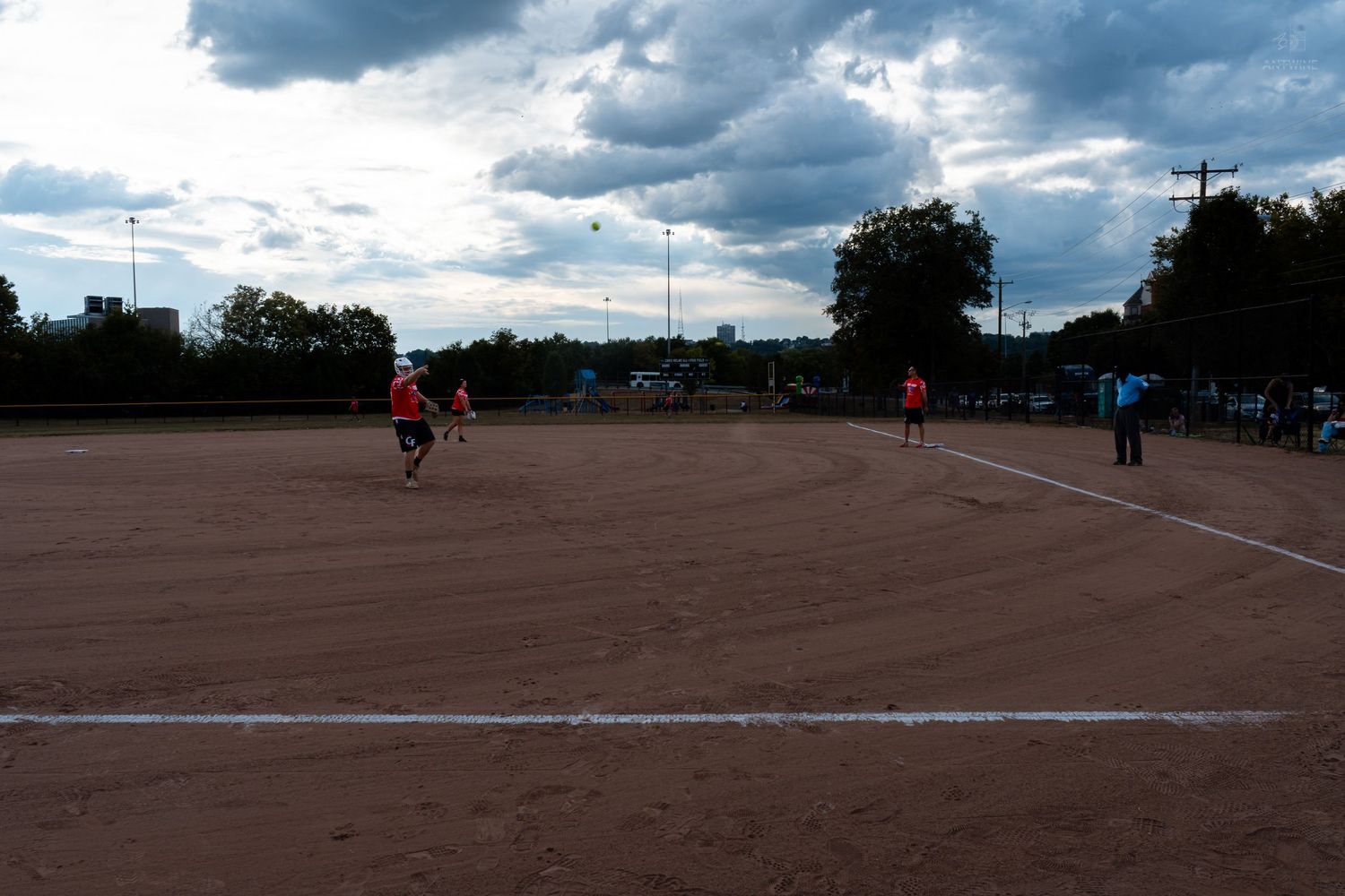Players in red uniforms compete on a dirt softball field under dramatic cloudy skies at dusk.