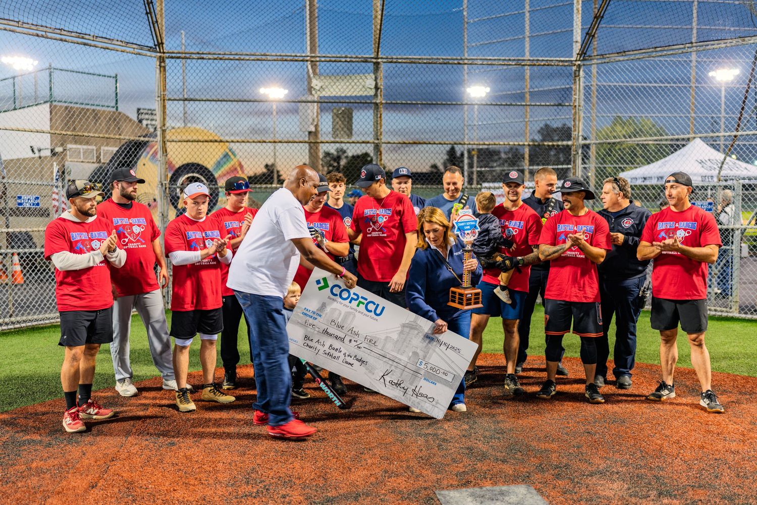 Winning softball team celebrates with oversized GAFCU check and trophy at night on baseball field.
