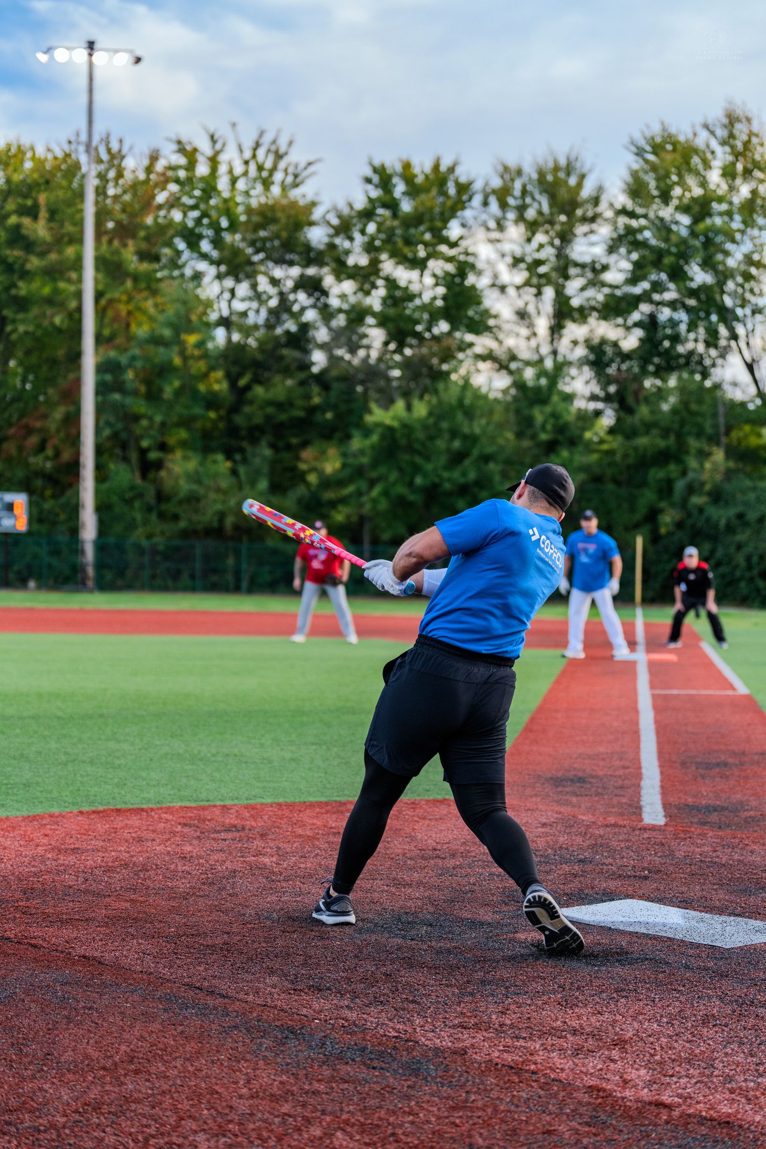 Softball batter in blue shirt follows through on swing during recreational game on red turf field.