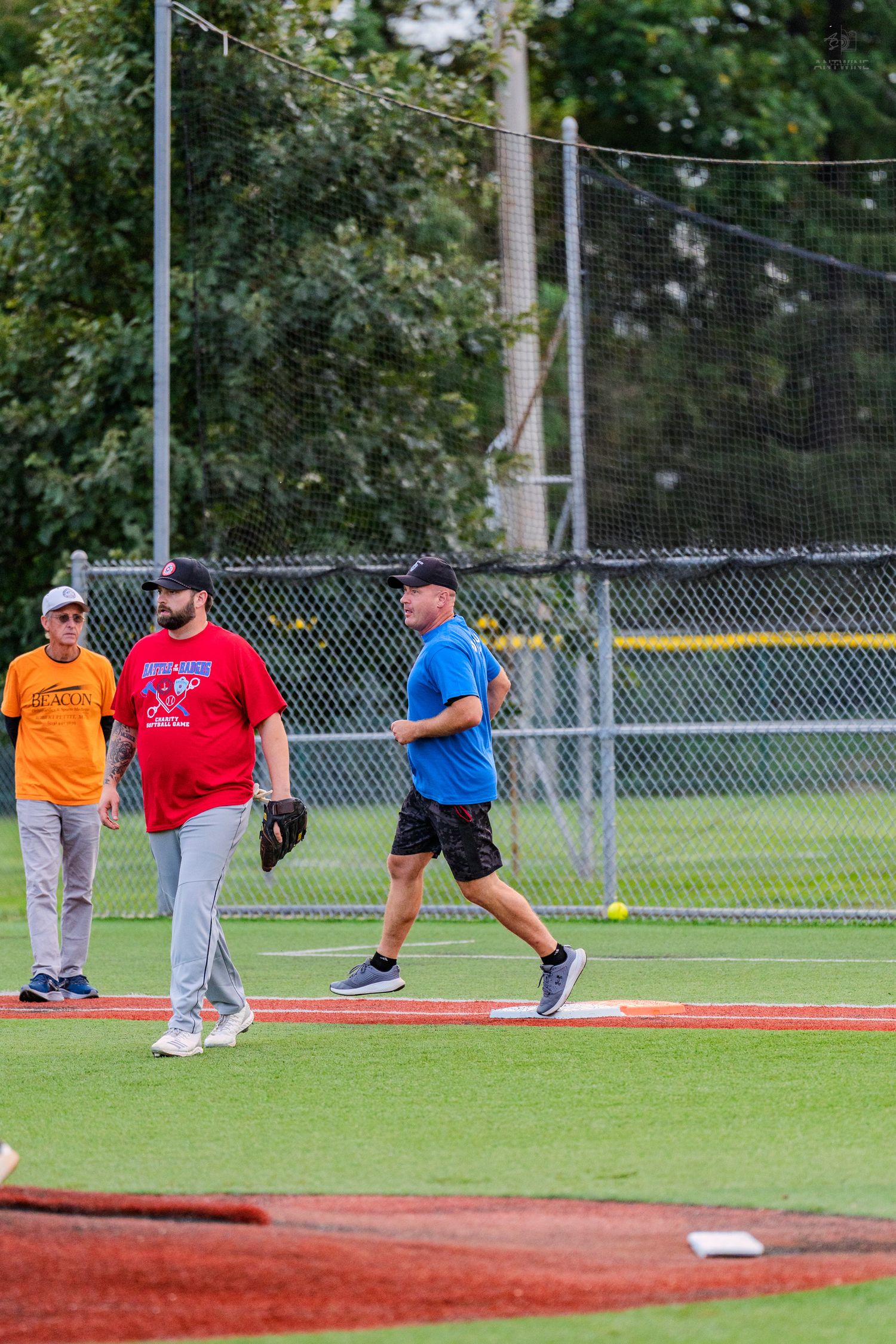 Three men playing baseball on a green field with a chain-link fence and trees in the background.