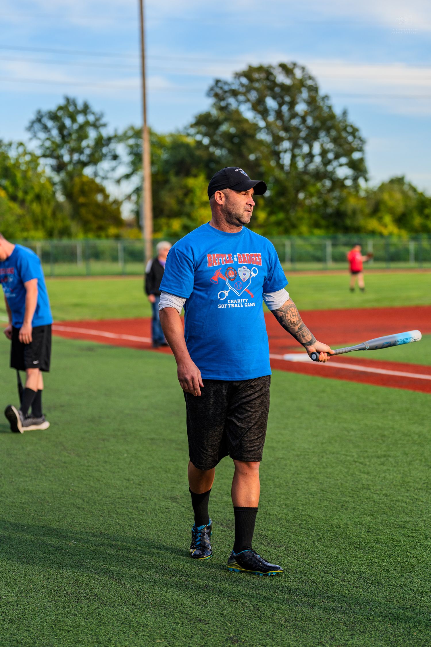 A tattooed man in a blue softball shirt holds a bat on a green turf baseball field.