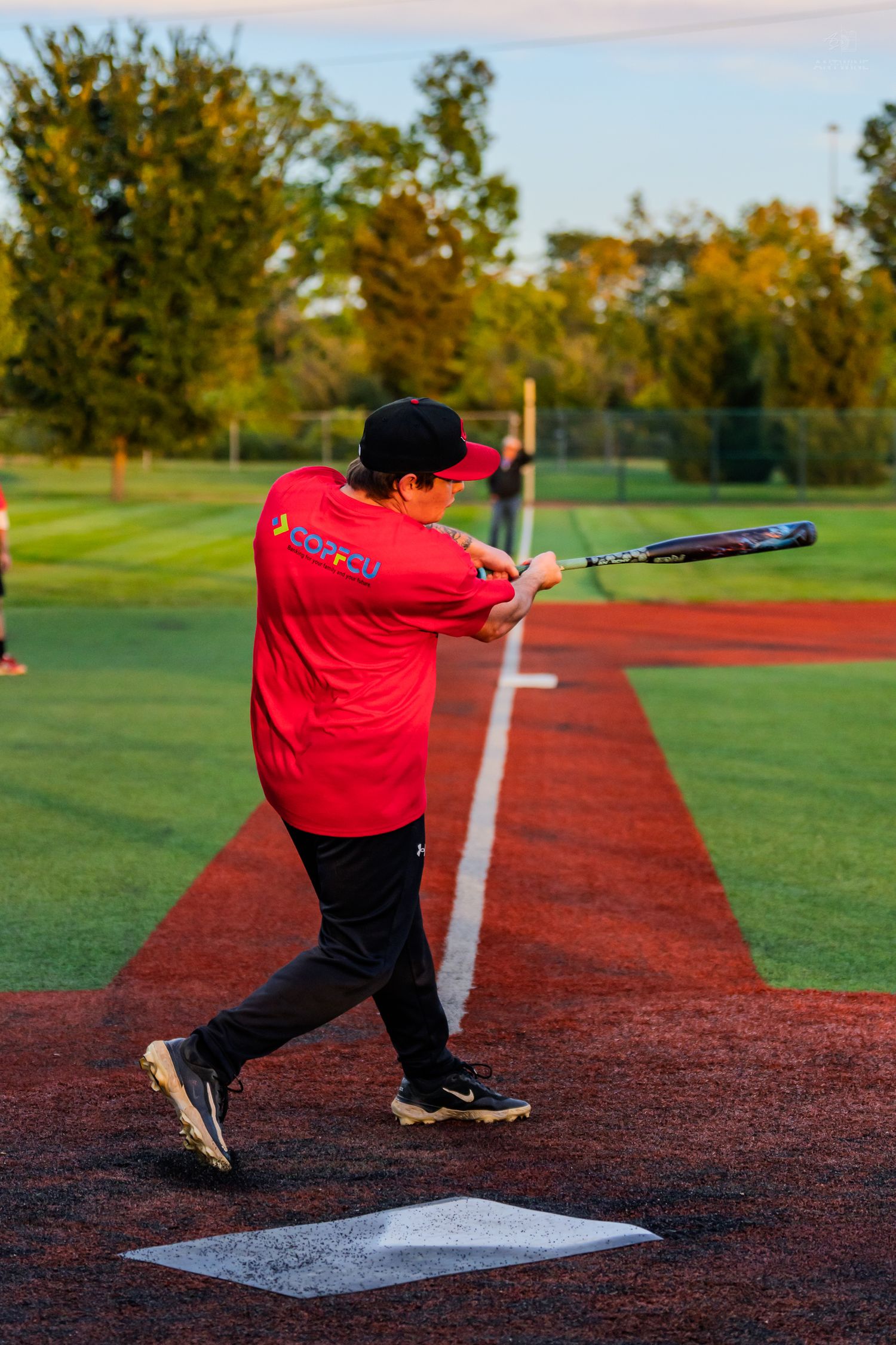 Baseball player in red shirt and black cap swinging bat at home plate on a baseball field at sunset.