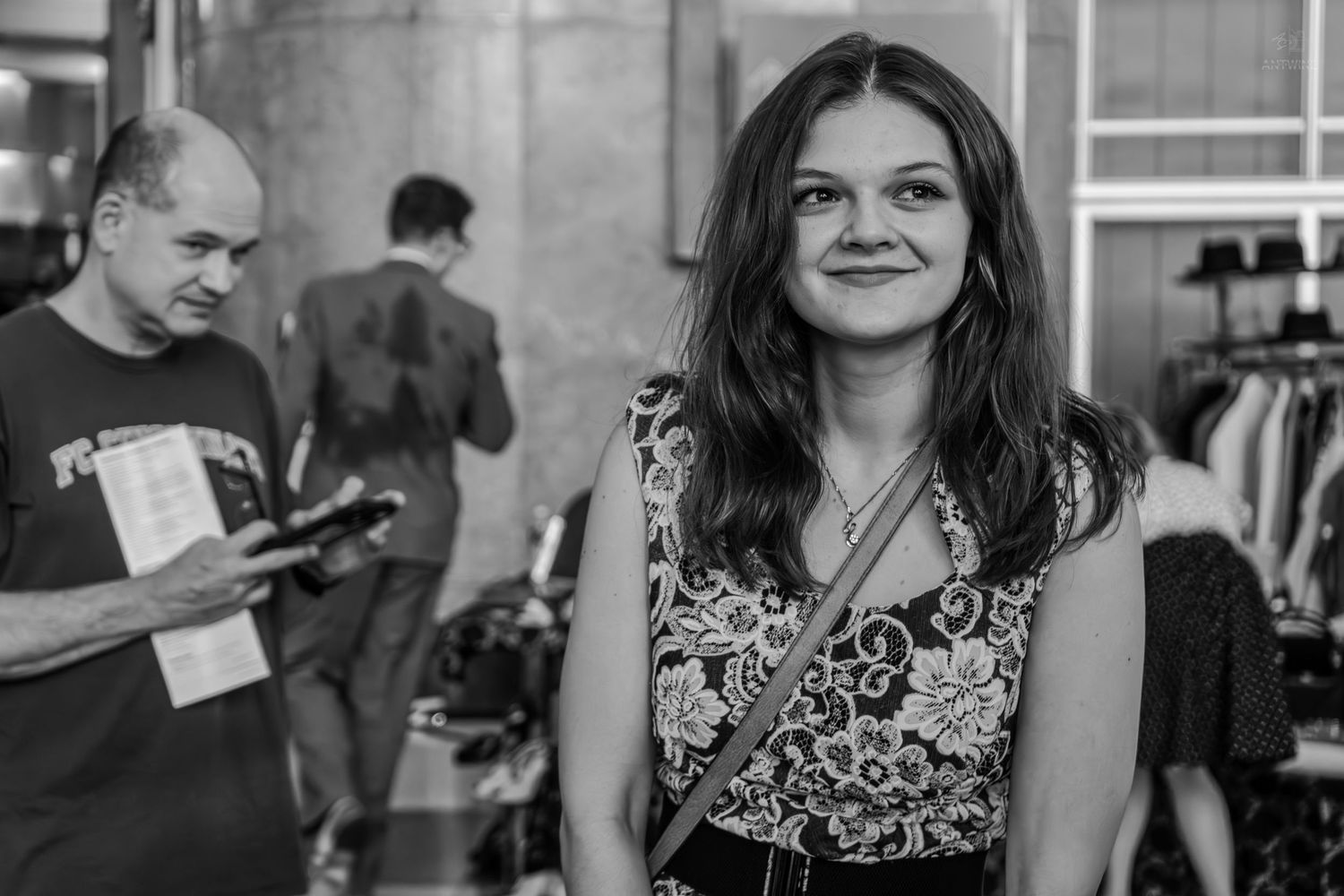Black and white photo of a smiling young woman at an indoor event, with people browsing in the background.