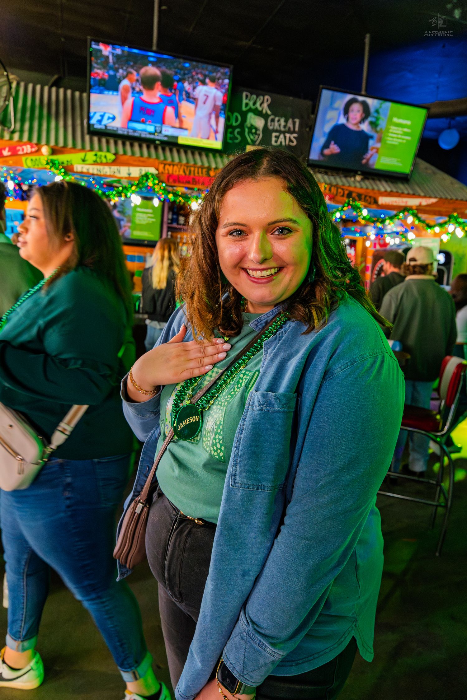Smiling woman in green top and blue jacket poses at a festive bar with TVs and colorful lights.