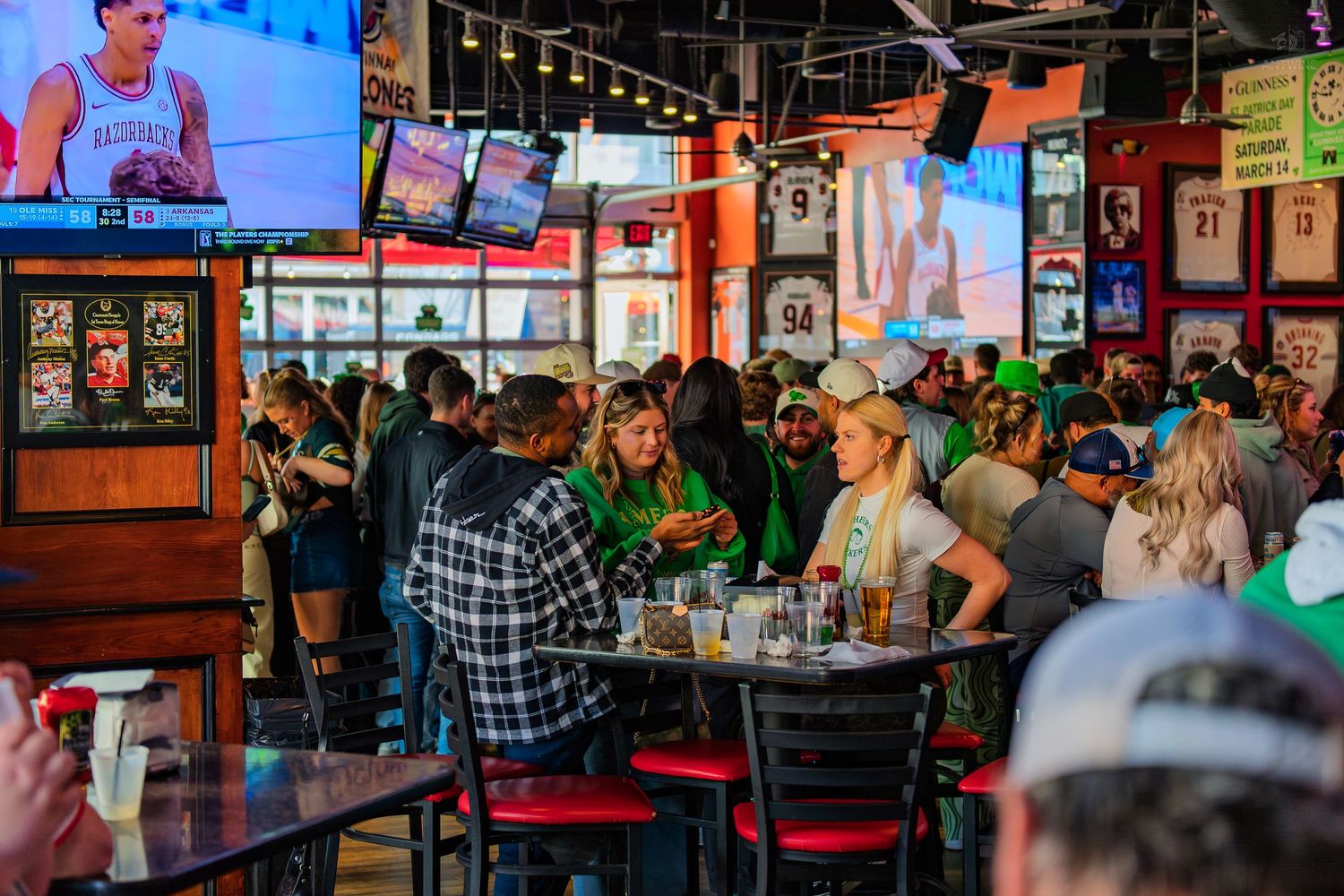 Crowded sports bar with fans watching games on multiple TVs, red chairs, and jerseys decorating the walls.