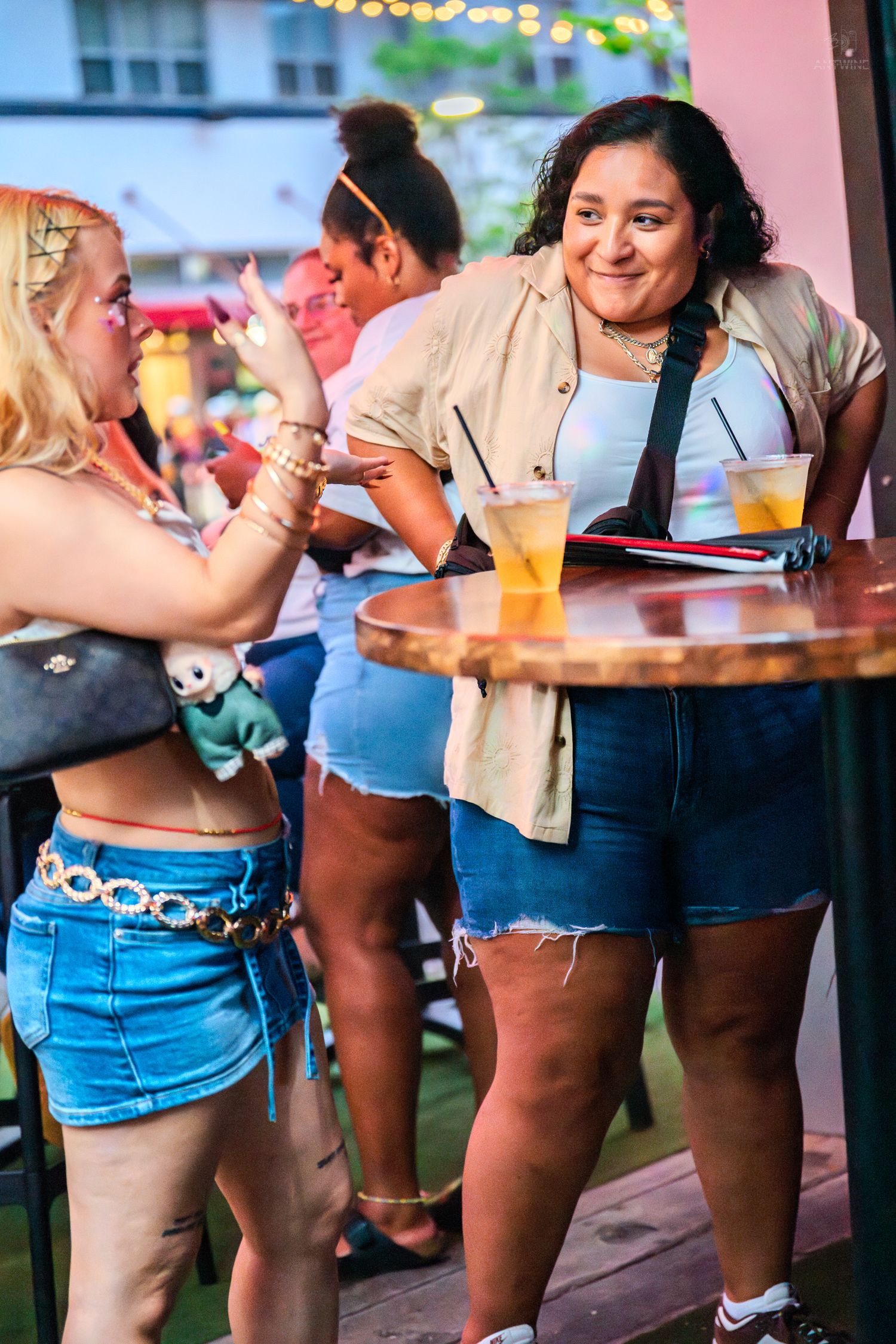 Women socializing at outdoor bar patio, holding colorful drinks, wearing denim shorts on warm summer evening.