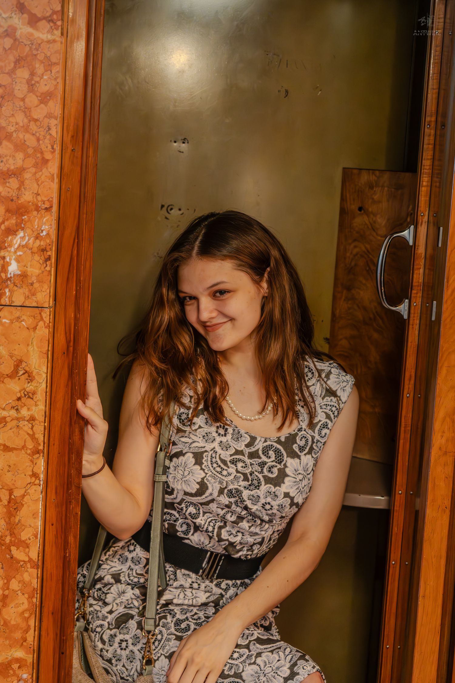 Young woman in floral dress smiling inside an ornate elevator with marble walls and wooden door frame.