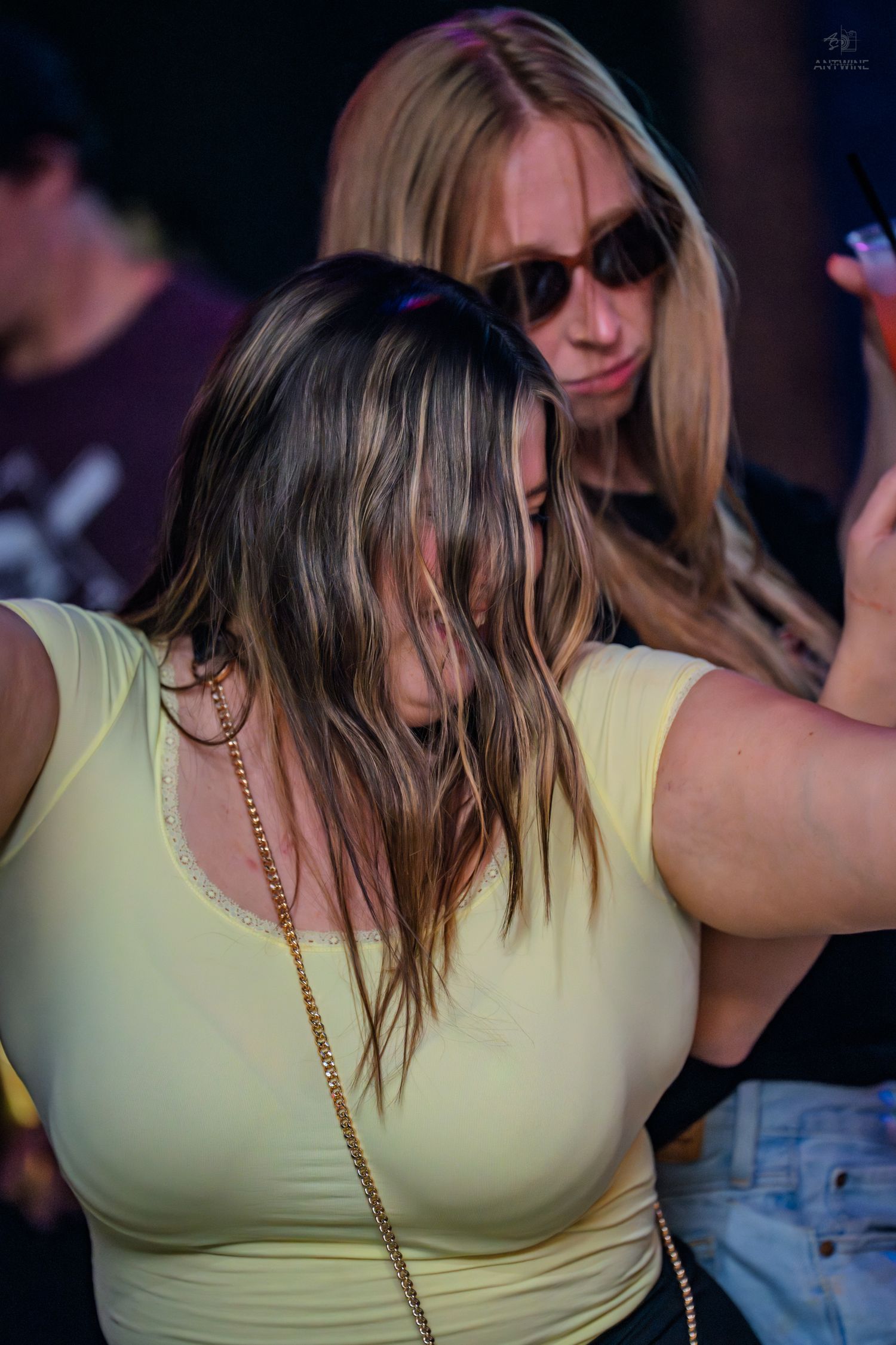 Two young women dancing at a nightclub event, one in a yellow crop top with a gold chain bag.
