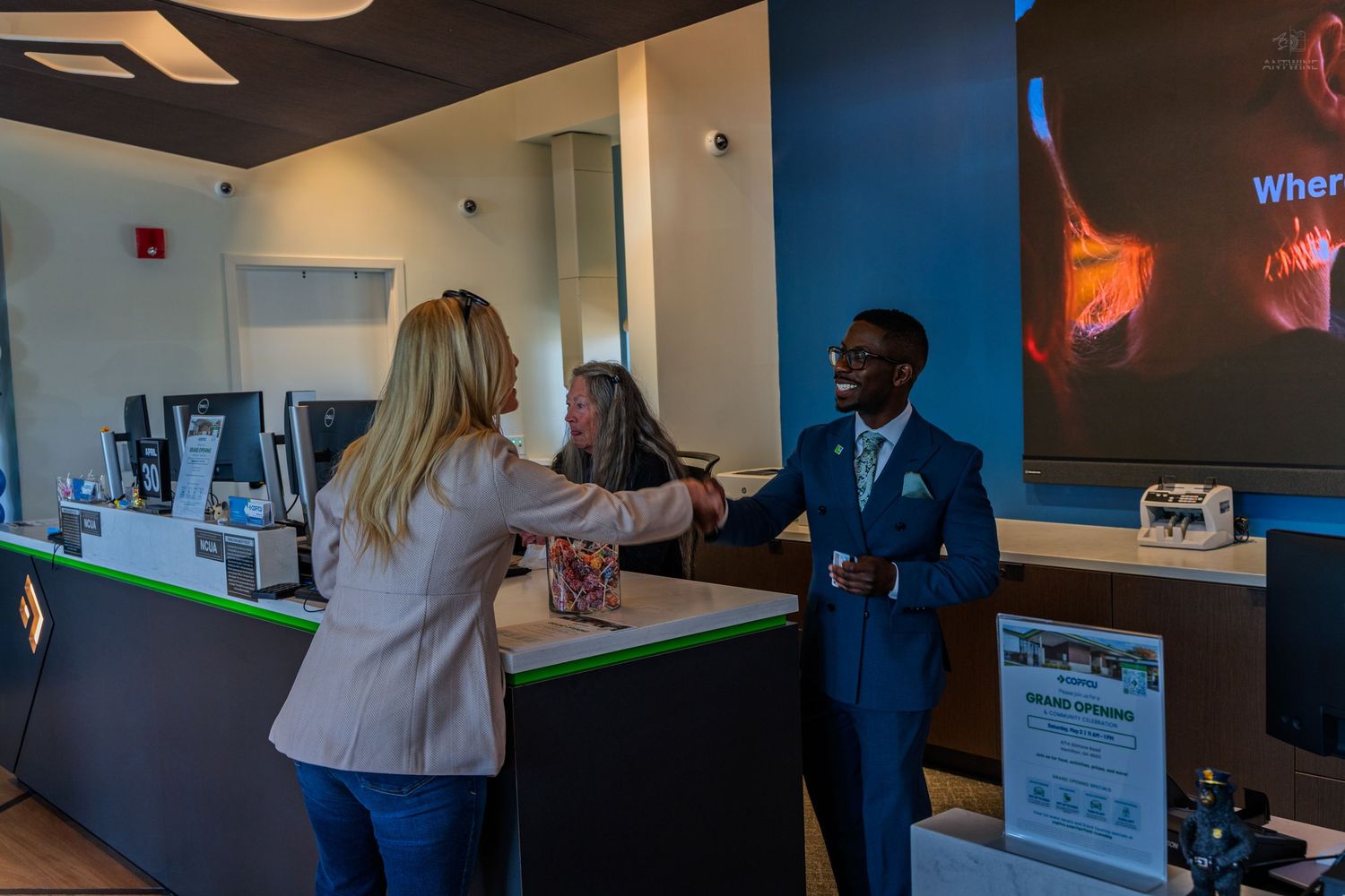 A COPFCU bank employee in a blue suit shakes hands with a customer at a modern credit union branch counter.