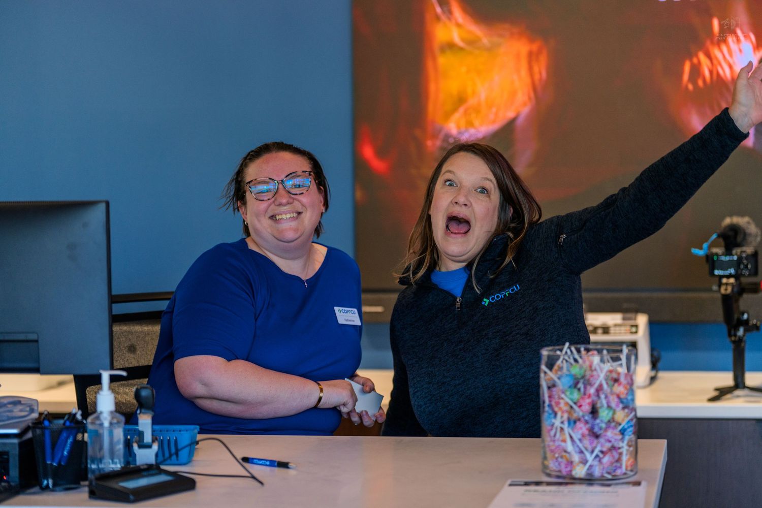 Two women smile and celebrate behind a counter in a blue-lit retail store, one raising her arm in excitement.