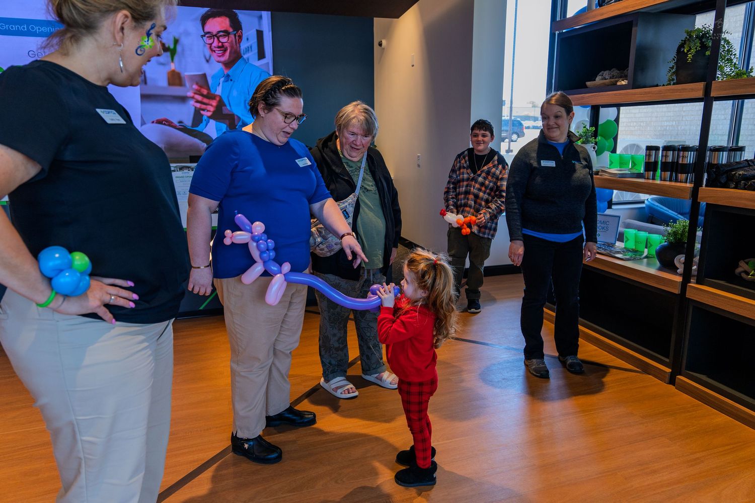 Staff and guests watch a toddler in red play with a balloon animal at an indoor children's event.