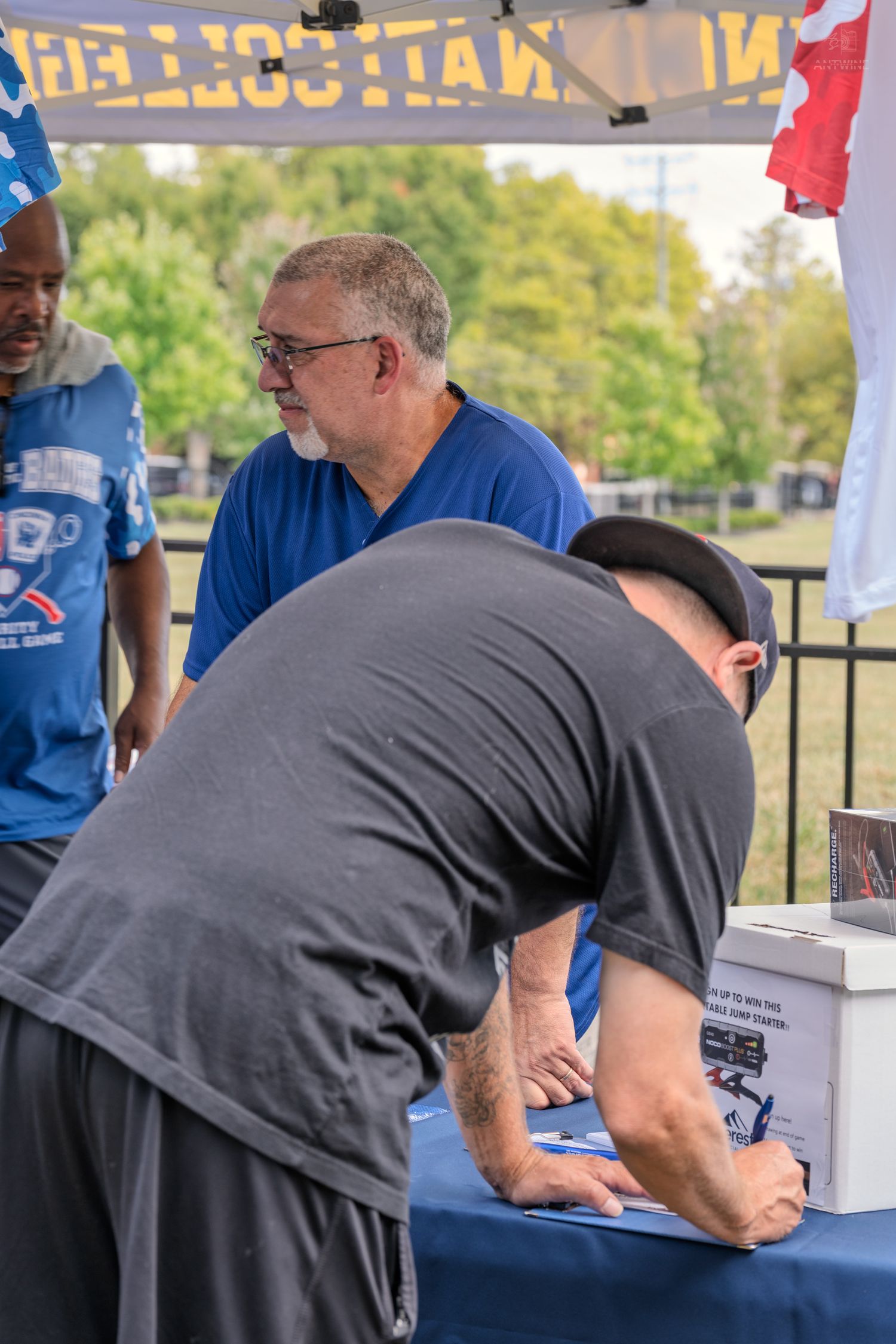 A man leans over a registration table at a charity softball game while an older man in blue watches.