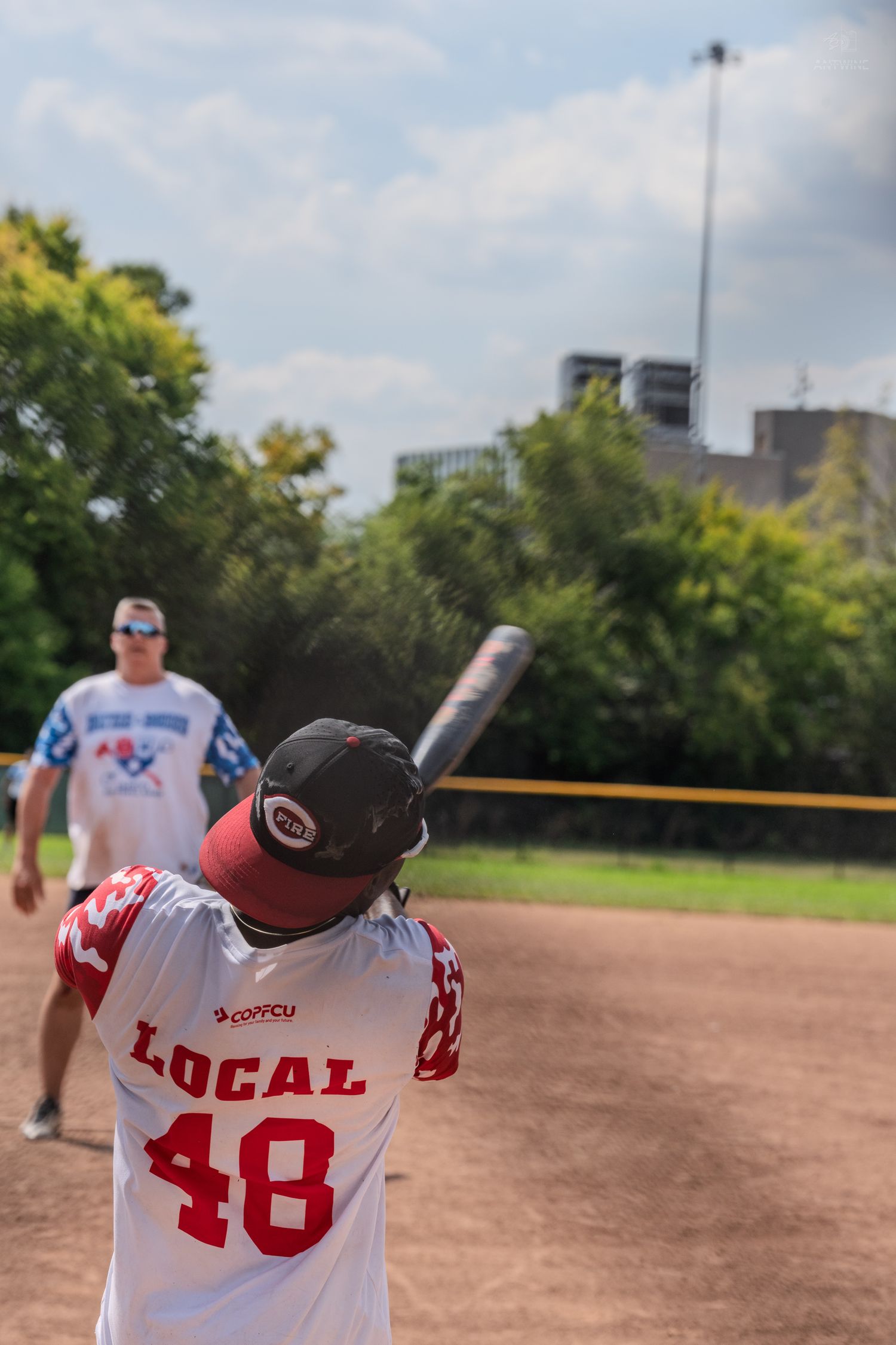 Batter in Local 48 jersey at plate preparing to swing during an outdoor softball game on a cloudy day.