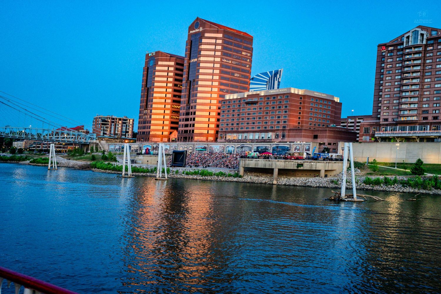 Columbus Ohio skyline reflecting on the Scioto River at dusk with illuminated buildings and a suspension bridge.