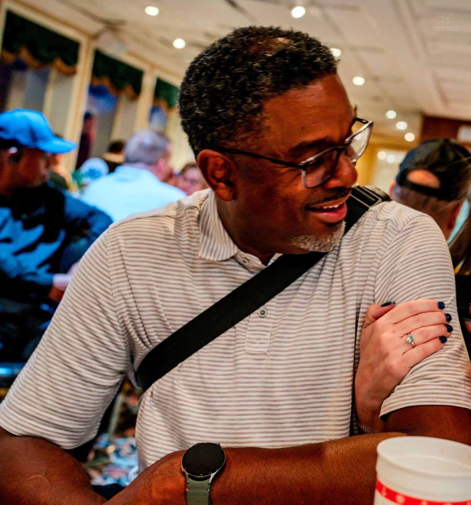 A man in a striped polo shirt wearing glasses and a crossbody bag smiles at an indoor gathering.