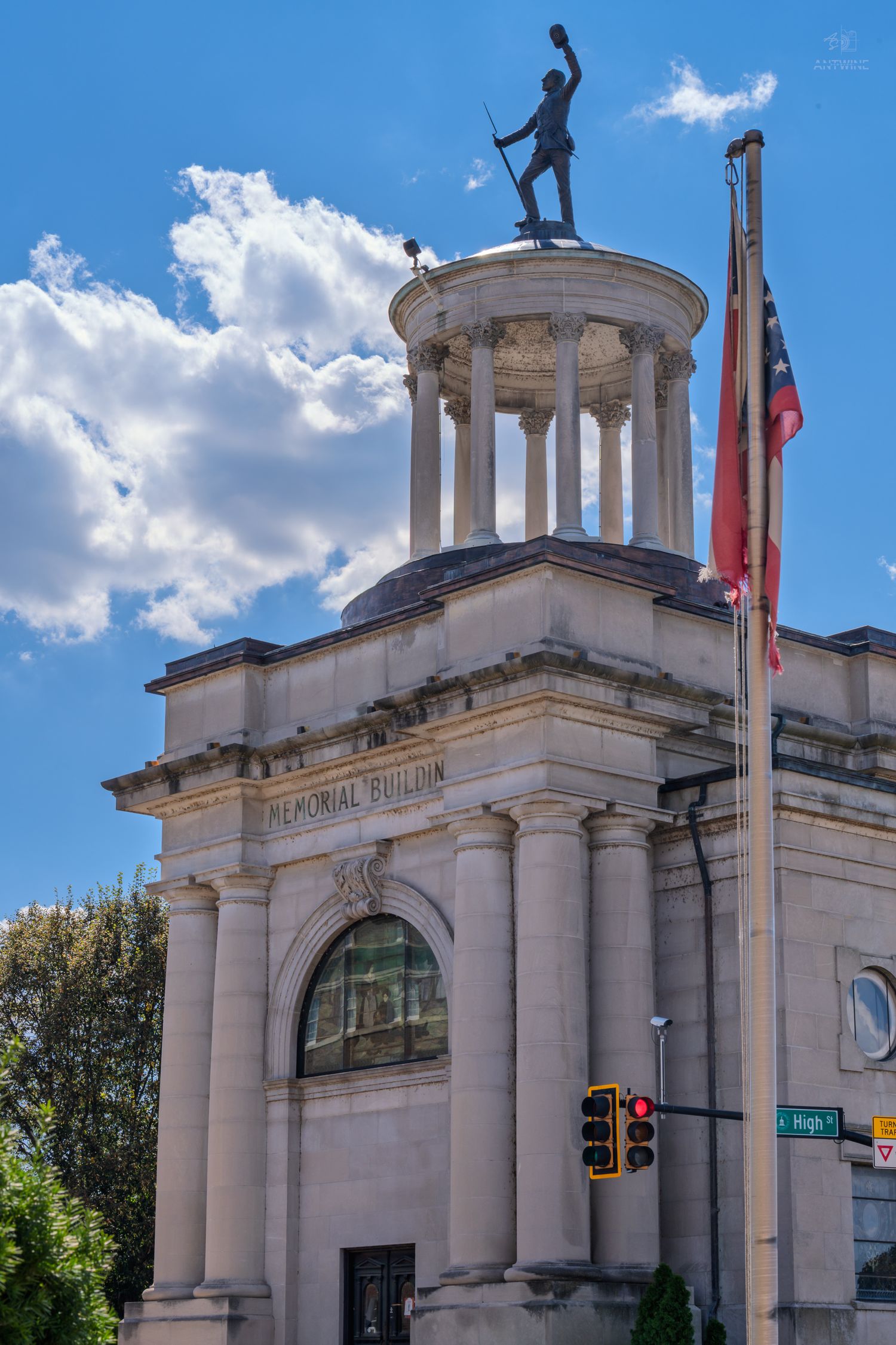 Historic Memorial Building with classical columns and soldier statue atop rotunda against blue sky with American flag.