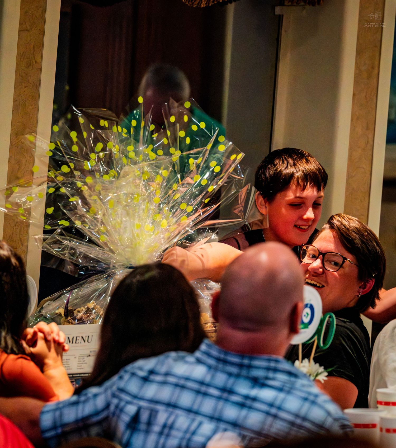 Guests at an indoor event gather around a large decorative gift basket with yellow ribbon accents.