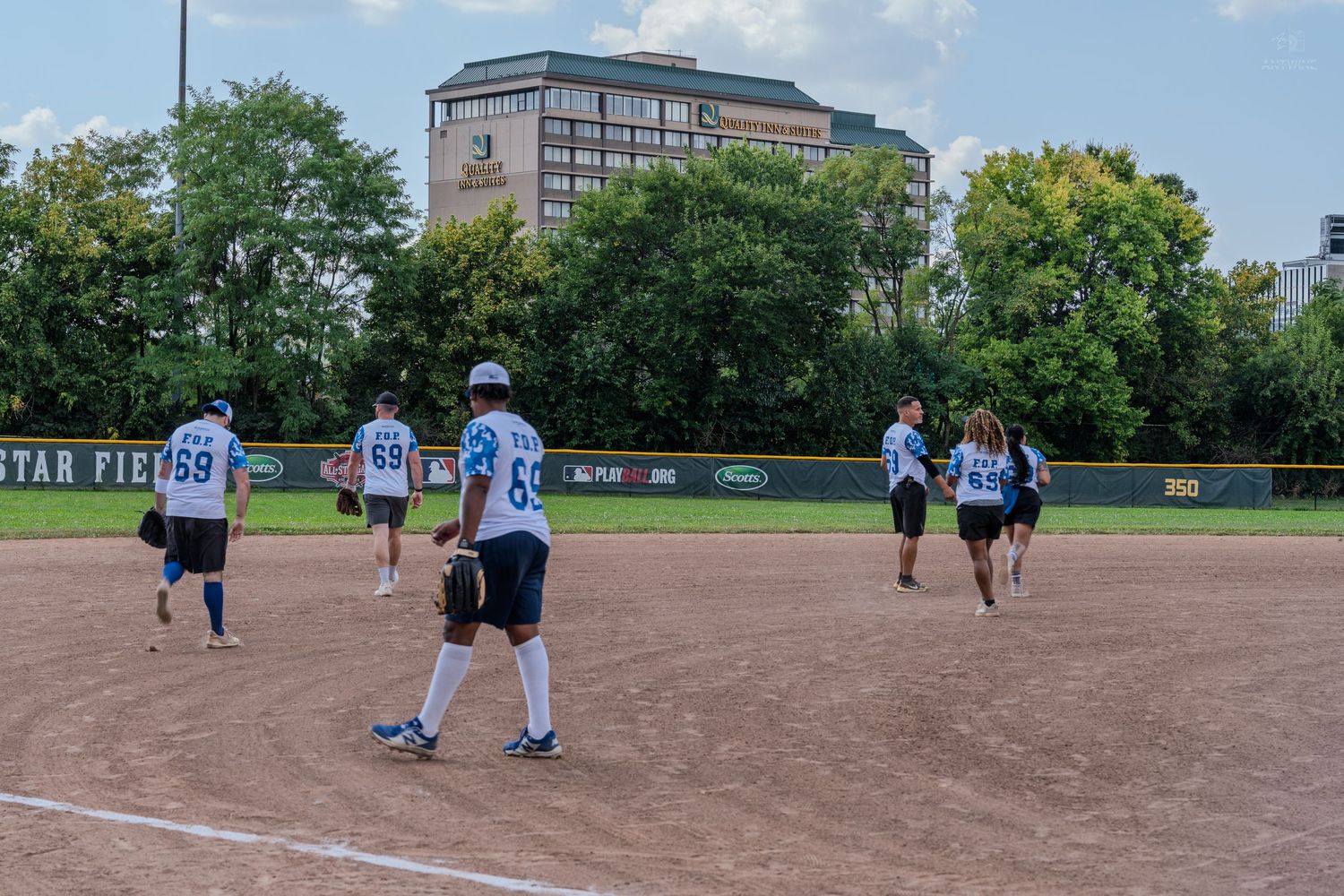 softball players on dirt field with building and trees in background during outdoor game.