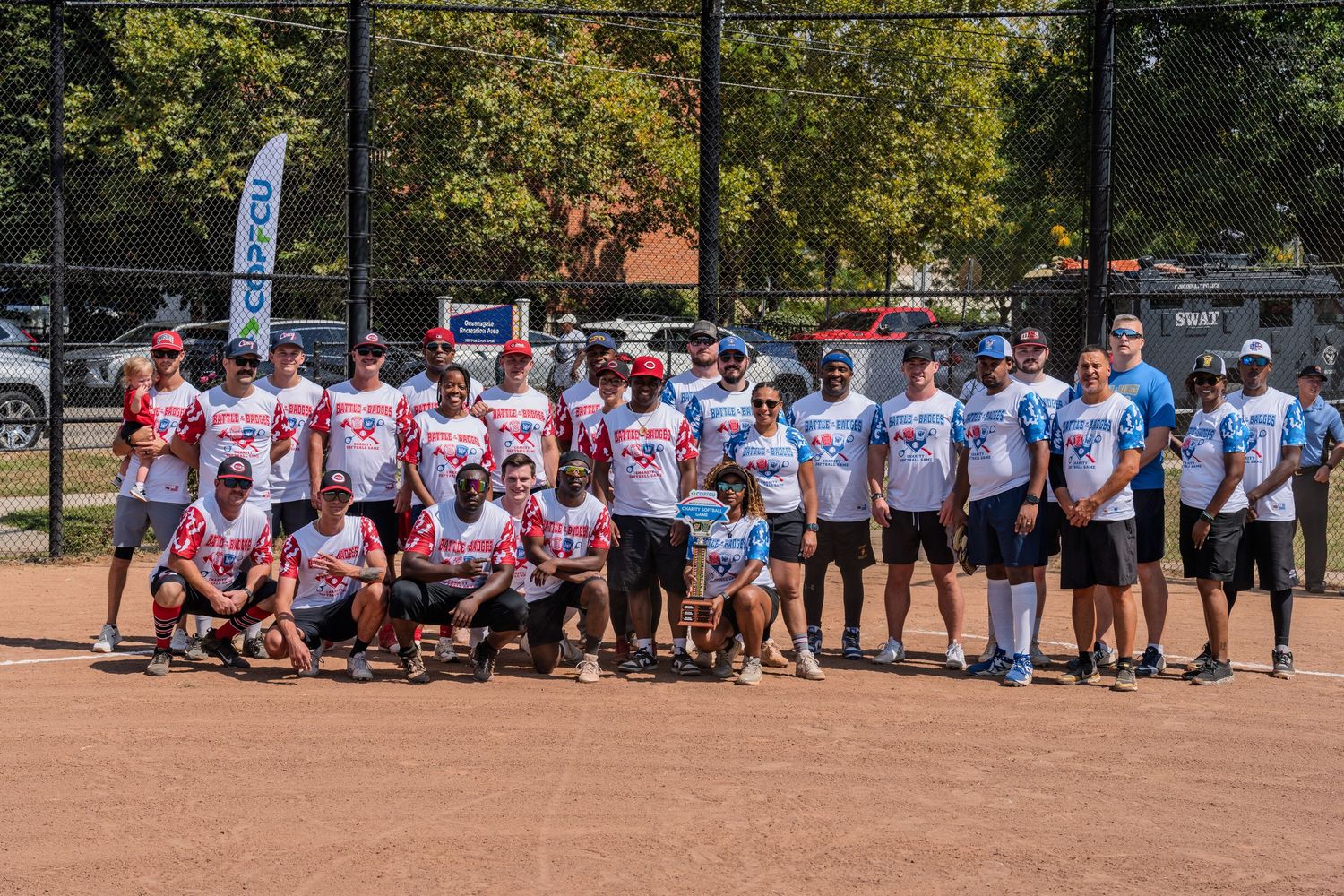 Large group of softball players posing together on a baseball diamond on a sunny day outdoors.