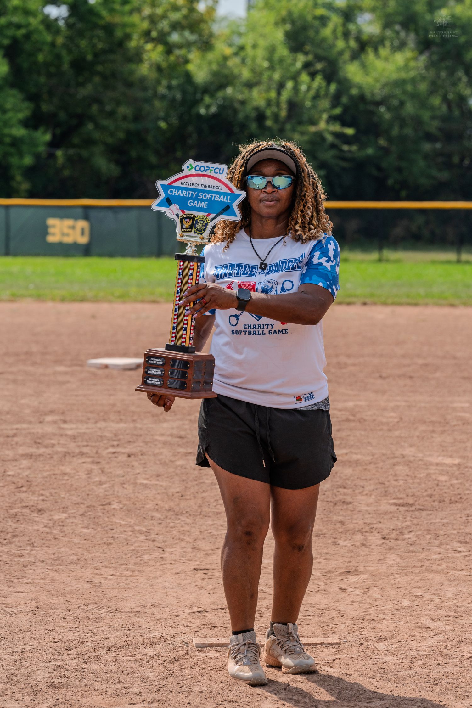 Woman holds a Charity Softball Game trophy on a baseball field on a sunny day.
