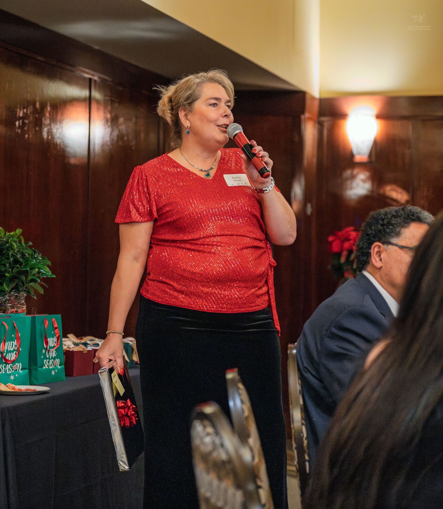 Woman in red sequin top speaks into microphone at festive holiday party event with guests seated nearby.