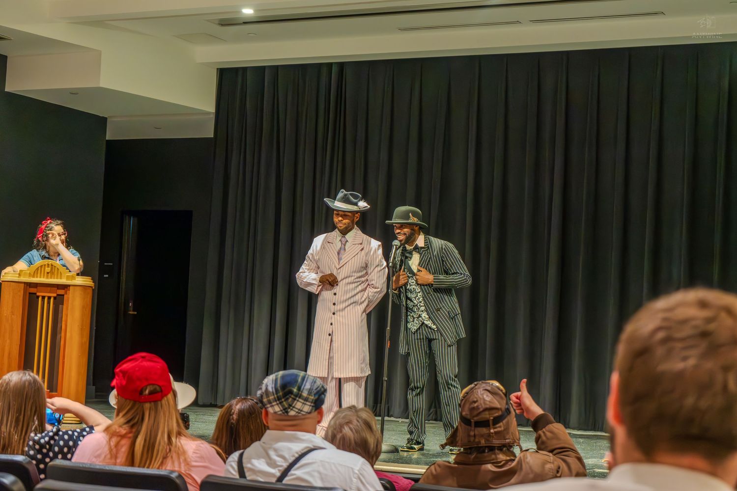 Two performers in vintage suits and hats on stage before an audience at an indoor theater event.