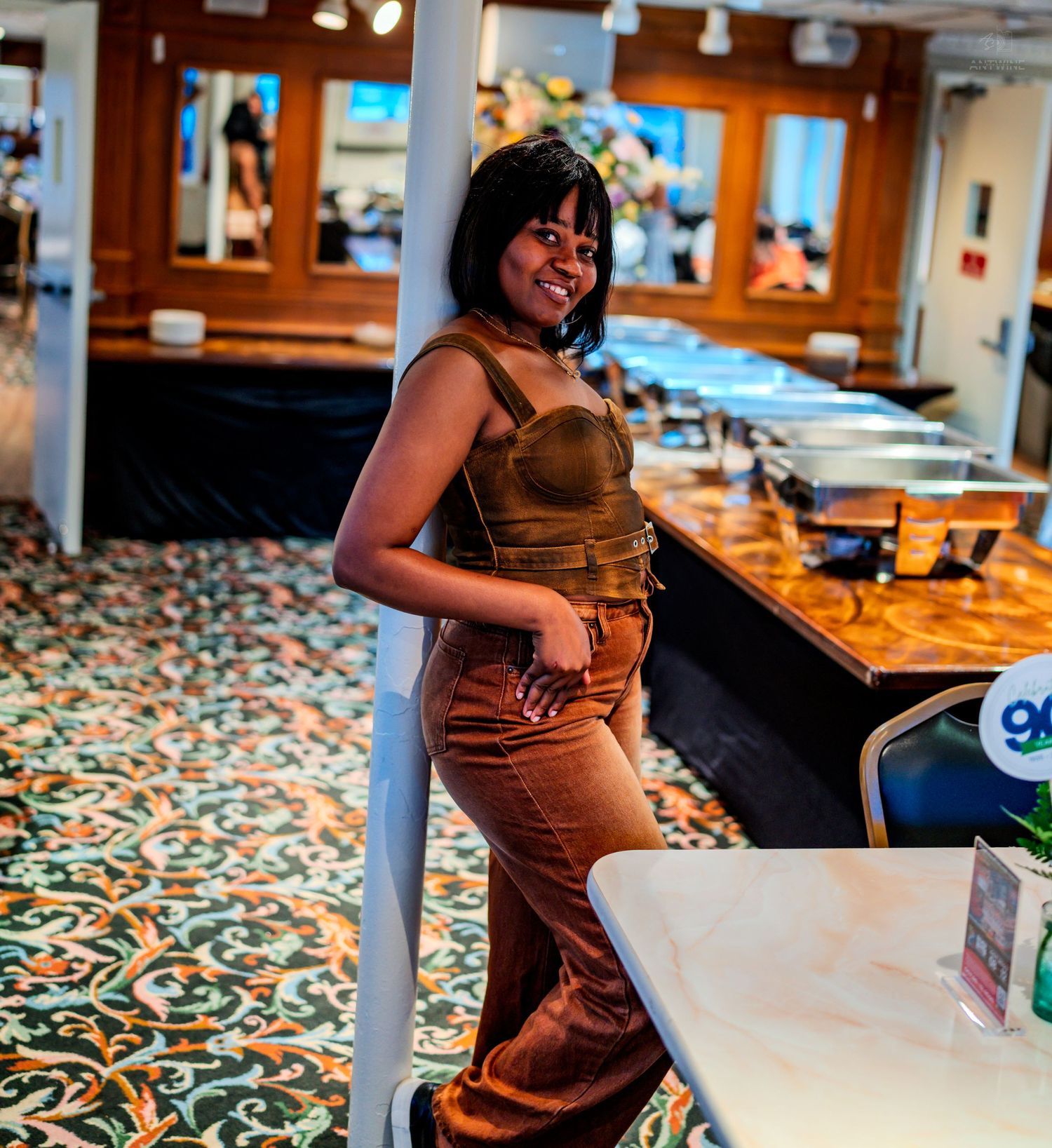 Woman in brown corset top and wide-leg pants posing confidently in a stylish restaurant interior.