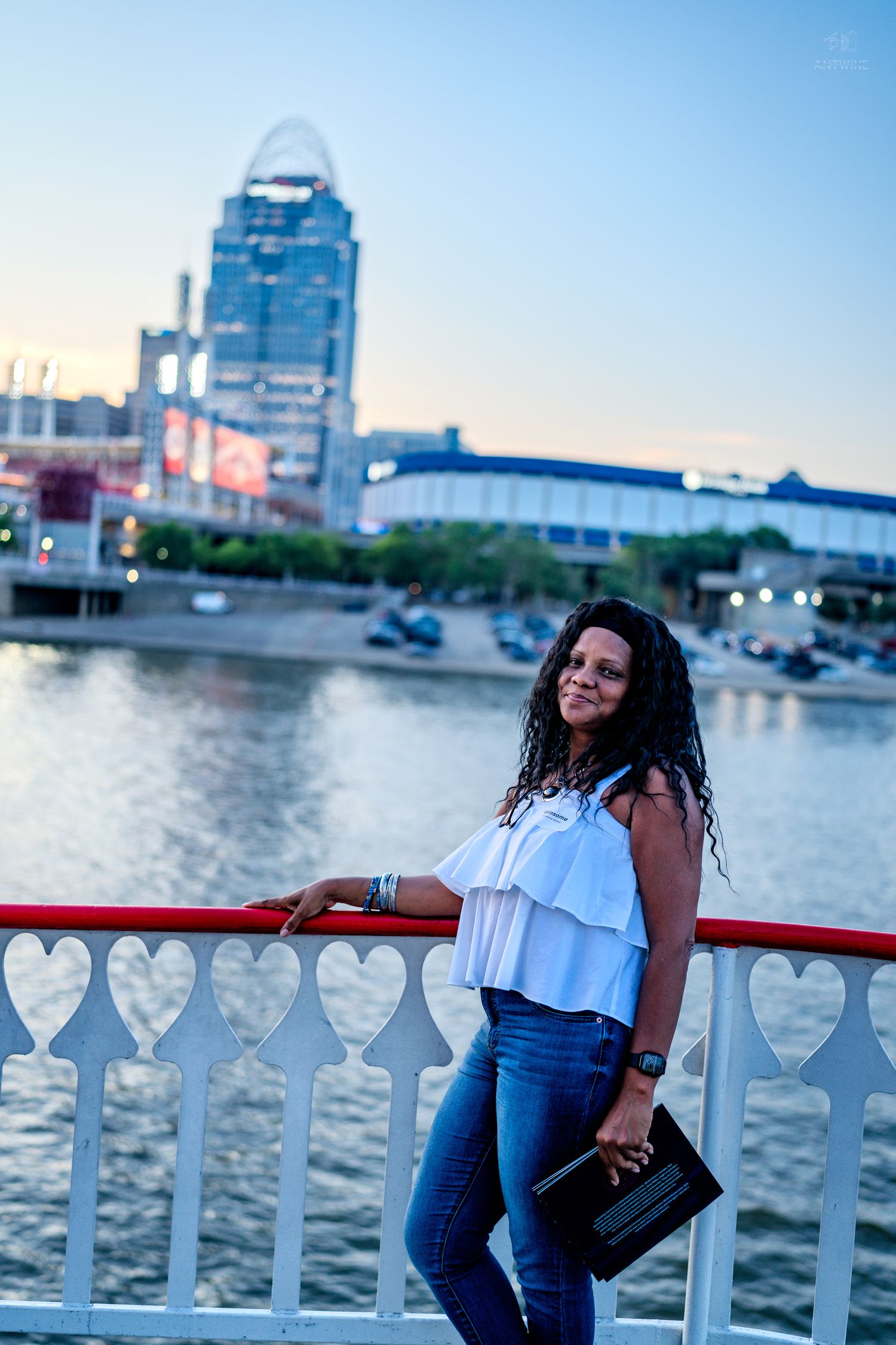 Woman in white top and jeans poses by red railing with Cincinnati skyline and Ohio River at dusk.