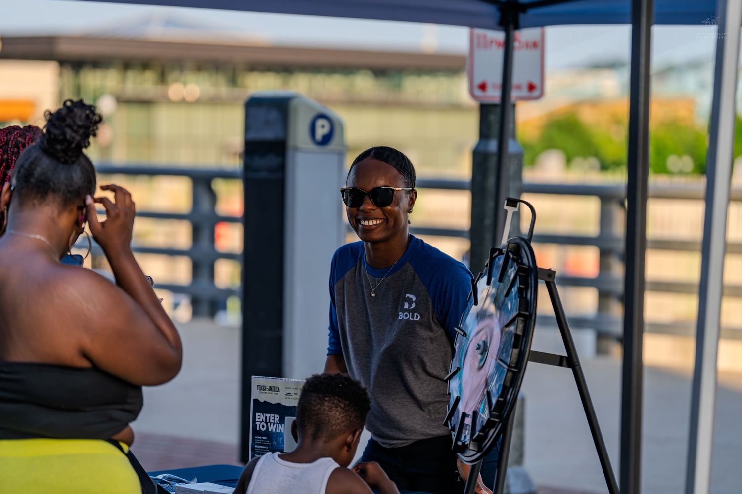 Woman smiling at an outdoor event under a canopy near a parking sign, with another woman and child nearby.