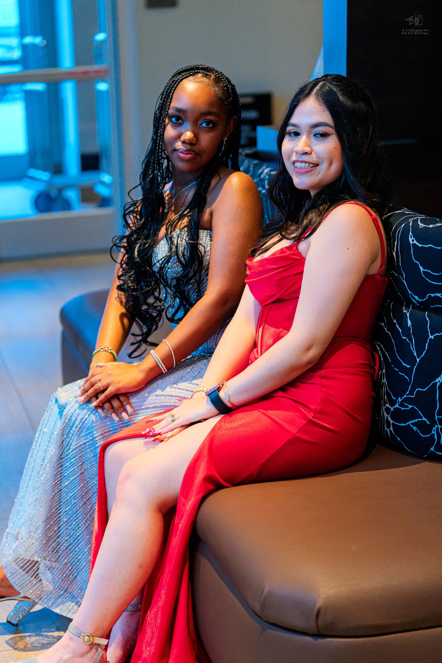 Two young women in elegant formal dresses, one silver and one red, sitting together smiling at a prom event.